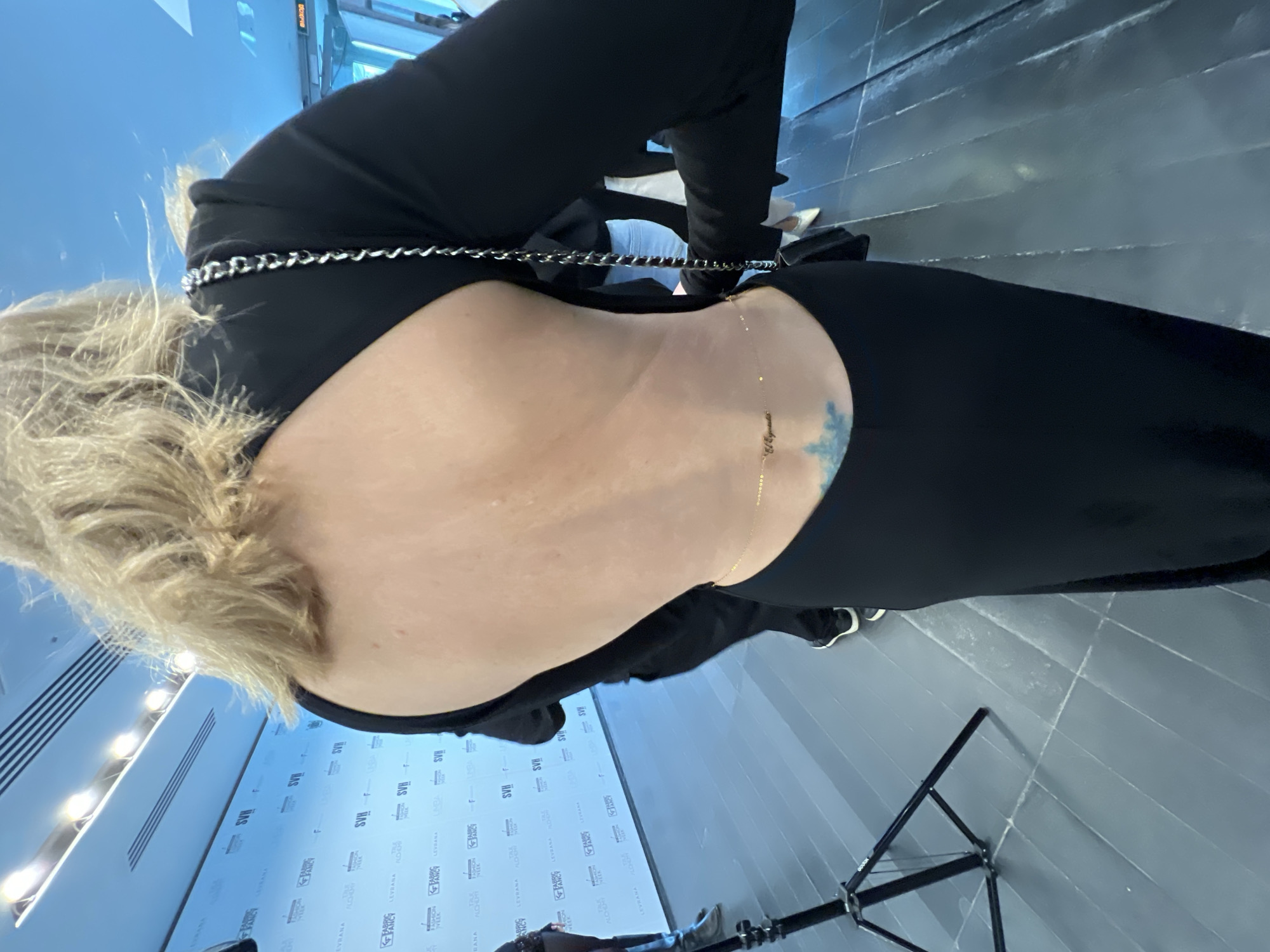 Here is a description of the image:
A woman with blond hair and wearing a black dress is seen from behind. The dress has a large cutout in the back, revealing most of her back. A thin gold chain drapes across her lower back, near a small tattoo on her right side. A silver chain appears to be part of the dress's design around the upper back.
The background shows a wall with logos and what appears to be promotional material, potentially for a fashion or beauty event. A photography light stand is visible in the lower right corner. The floor is a dark grey or black color with diagonal lines.