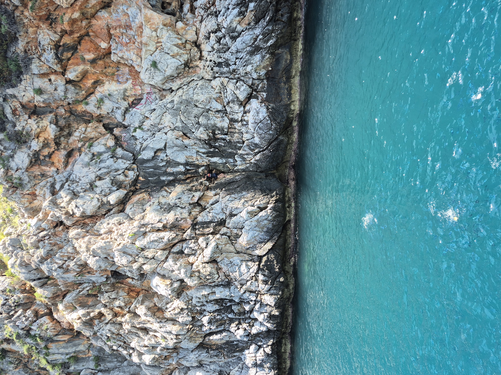 An aerial shot shows a rocky cliff face meeting the turquoise water of the sea. Two people are positioned at the cliff base. The shot is taken at Alanya, Turkey.