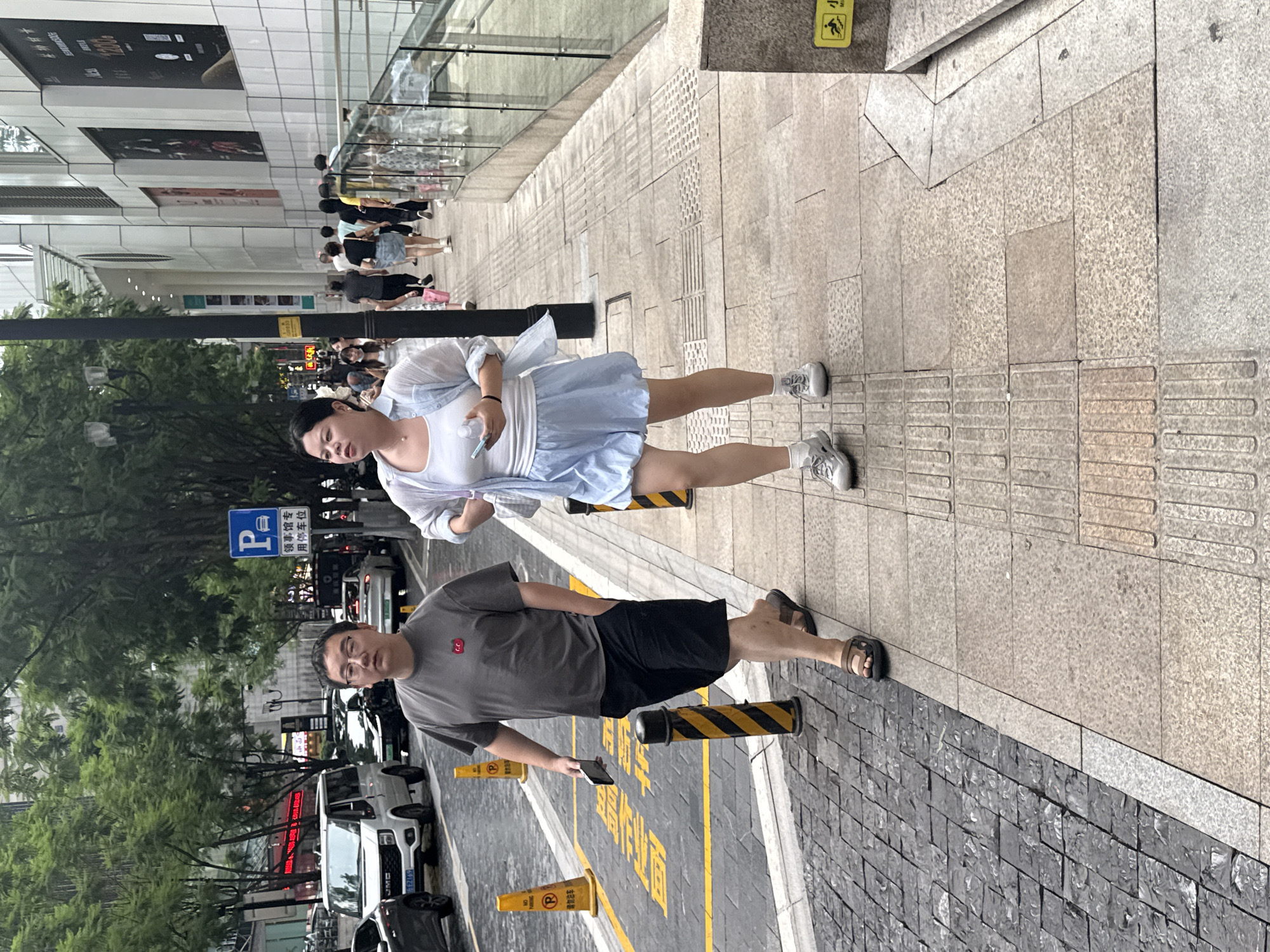 In Yuzhong District, China, on a sidewalk next to a street with vehicles and trees, a man and a woman are walking side-by-side. The man, wearing a grey t-shirt and black shorts, holds a phone in his right hand and has his left hand in his pocket. The woman, in a white tank top and light blue shorts, carries a phone and a bottle in her hands and wears a light blue unbuttoned shirt over her tank top. They are walking on a sidewalk composed of textured and smooth paving stones, bordered by a street with yellow lines and traffic cones. In the background, a white van, other cars, and pedestrians are visible, along with a modern building facade and trees.