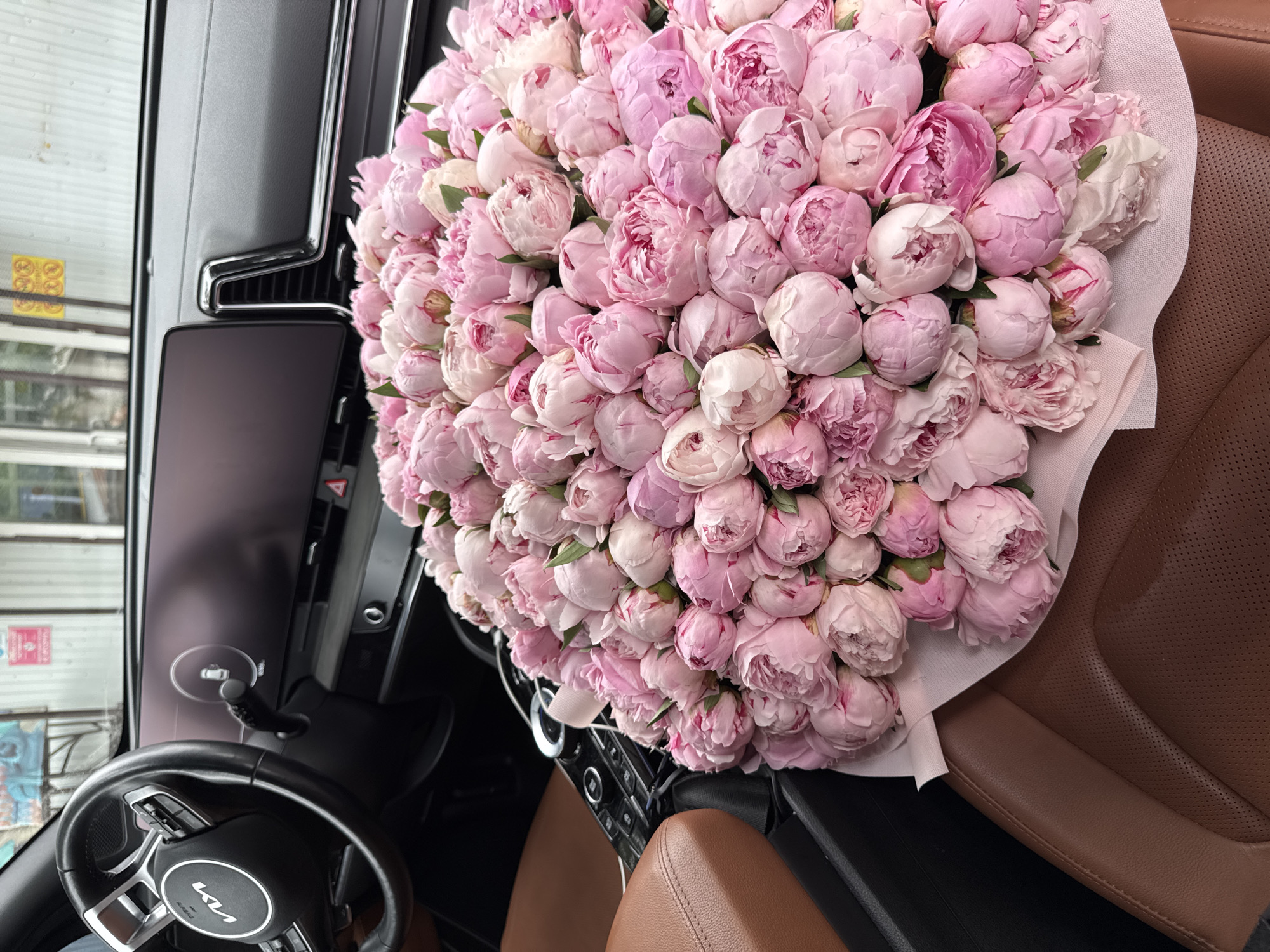 The image shows a large bouquet of pink peonies placed on the passenger seat of a car. The car's interior is visible, including the steering wheel with the "KIA" logo, the dashboard with a digital display, and brown leather seats. The background suggests an urban environment, possibly a street or parking area, with some graffiti on a wall. The lighting is bright, indicating daytime. There are no people visible in the image. The scene appears to be set in Almaty, Kazakhstan, based on the provided context.