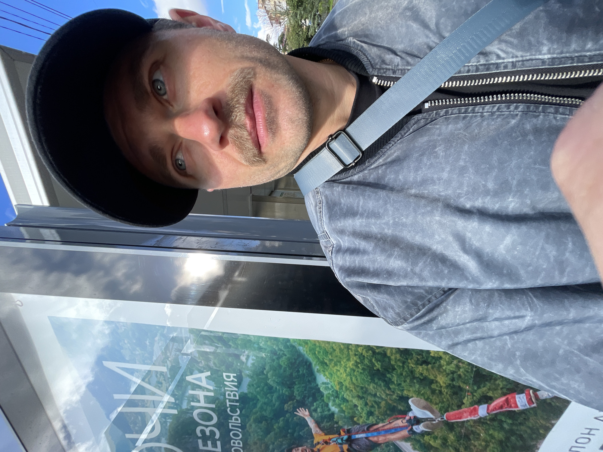 A man wearing a black baseball cap and gray jacket is posing for a selfie in front of a poster. The poster is for "Skypark" Sochi Russia, featuring a person doing a bungee jump with the slogan "Season of Pleasures". The man is wearing a gray shoulder bag. The background appears to be a city street with buildings and trees. The overall lighting is bright, suggesting it is daytime.
