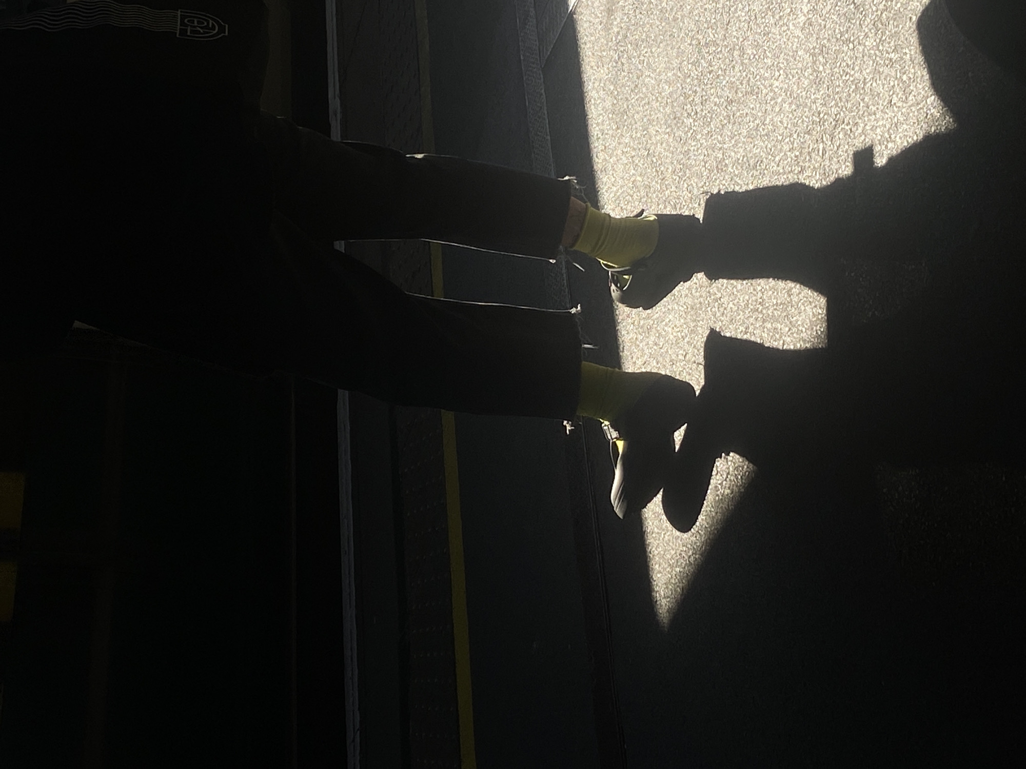 This image, taken in London, United Kingdom, captures a person standing or walking between a deeply shadowed area and a brightly sunlit path. The strong, direct sunlight creates a dramatic contrast, casting a prominent, long shadow of the individual onto the textured, light-colored ground, likely concrete or pavement.
The subject is partially visible, primarily silhouetted against the dark background. They are wearing dark trousers or jeans, which appear slightly distressed near the ankles, vibrant yellow socks, and light-colored sneakers. A logo is faintly visible on the upper back of their dark clothing in the shadowed area, depicting wavy lines leading to a stylized letter "P" enclosed within a circular or oval shape.
The shadowed environment appears to be an interior space, a covered walkway, or an underpass, characterized by dark surfaces. A distinct vertical yellow line is visible on a dark floor or wall in this shadowed section, possibly a safety marking. The bright illumination of the ground indicates a sunny day.
The image focuses on the interplay of light and shadow, highlighting the person's outline and the bright accents of their socks against the stark background.