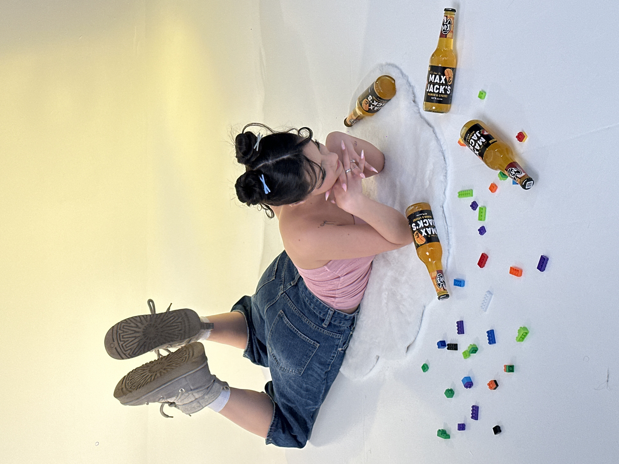 The image depicts a woman with dark hair styled in buns, secured with blue hair clips, kneeling on a white surface, likely a faux fur rug, surrounded by bottles labeled "MAX JACK'S MANGO & LYCHEE" and scattered colorful plastic building blocks. The woman is wearing a pink top and blue denim shorts. She's positioned with her back slightly turned, and her hands are near her face. The bottles are arranged around her, and the building blocks are dispersed on the white surface. She is wearing Ugg-like boots. The backdrop transitions from white to yellow. There is no indication in the image that it was taken in Yerevan, Armenia.