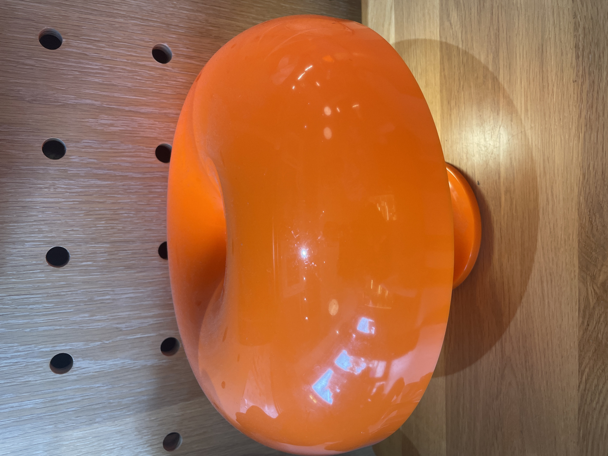The image displays a prominent, glossy, bright orange object set against a wooden background. The object is large, curvilinear, and appears to be a rounded, hollow form, possibly toroid or C-shaped, protruding from the wall. Its surface is highly reflective, showing various bright white and light blue highlights from ambient light sources, along with subtle reflections of the surrounding environment.
To the left of the orange object is a light-colored wooden panel, possibly a pegboard, featuring several uniformly spaced dark circular holes. This panel has a visible wood grain pattern running horizontally. To the right of the orange object is another section of a light-colored wooden surface, which also exhibits a wood grain. The orange object casts a soft, crescent-shaped shadow onto this right-hand wooden surface, indicating it is mounted or affixed to the wall.
There is no activity or event taking place; the image is a static shot of the object in its setting. The lighting suggests an indoor environment, likely under artificial illumination, given the distinct reflections. No people are visible, and there is no discernible text in the image. The scene appears to be an interior space, potentially in Bac Nha Trang Ward, Vietnam, showcasing a piece of modern design or functional art.