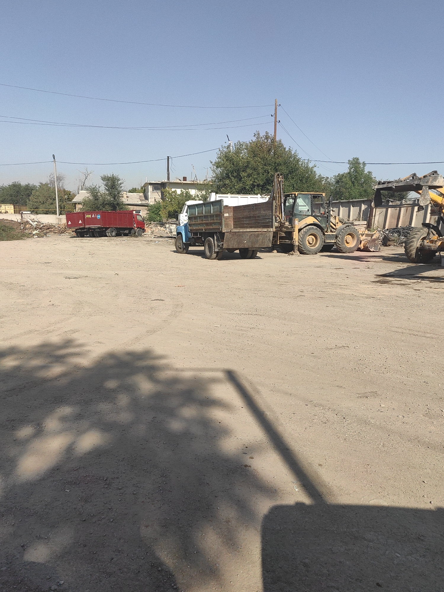 A medium-shot, outdoor photograph taken on a sunny day shows a dirt lot with several vehicles. A red truck with a long, silver-colored trailer is parked on the left side of the frame. In the center, a blue and white truck is parked, with its bed tilted upwards. Behind it, a yellow backhoe loader is positioned, and to its right, another piece of heavy machinery, possibly another excavator, is visible. The background consists of a few buildings and trees, suggesting an industrial or construction area. The ground is covered in gravel and dirt, with shadows cast by the vehicles and foliage. The location is identified as the city of Shymkent, Kazakhstan.
