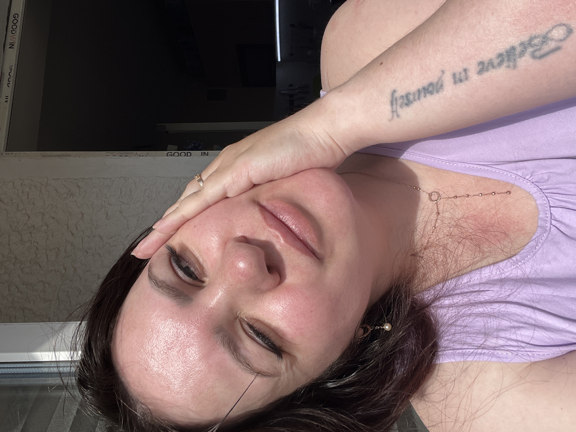 A woman is visible from the chest up, with her face tilted slightly towards the viewer, propped up by her right hand. The woman has dark hair, light skin, and closed eyes with eyeliner. Her lips are full and pursed. She wears a lilac-colored top and a gold necklace with a circular charm. A small earring with a pearl is visible in her right ear. On her right forearm, the tattoo "Believe in yourself" is visible. The background shows a window frame with the text "GOODWIN" visible. Behind the window, a dark room or outdoor area is discernible. The location is not possible to identify as the city of Noviy, Russia, based on the provided information.