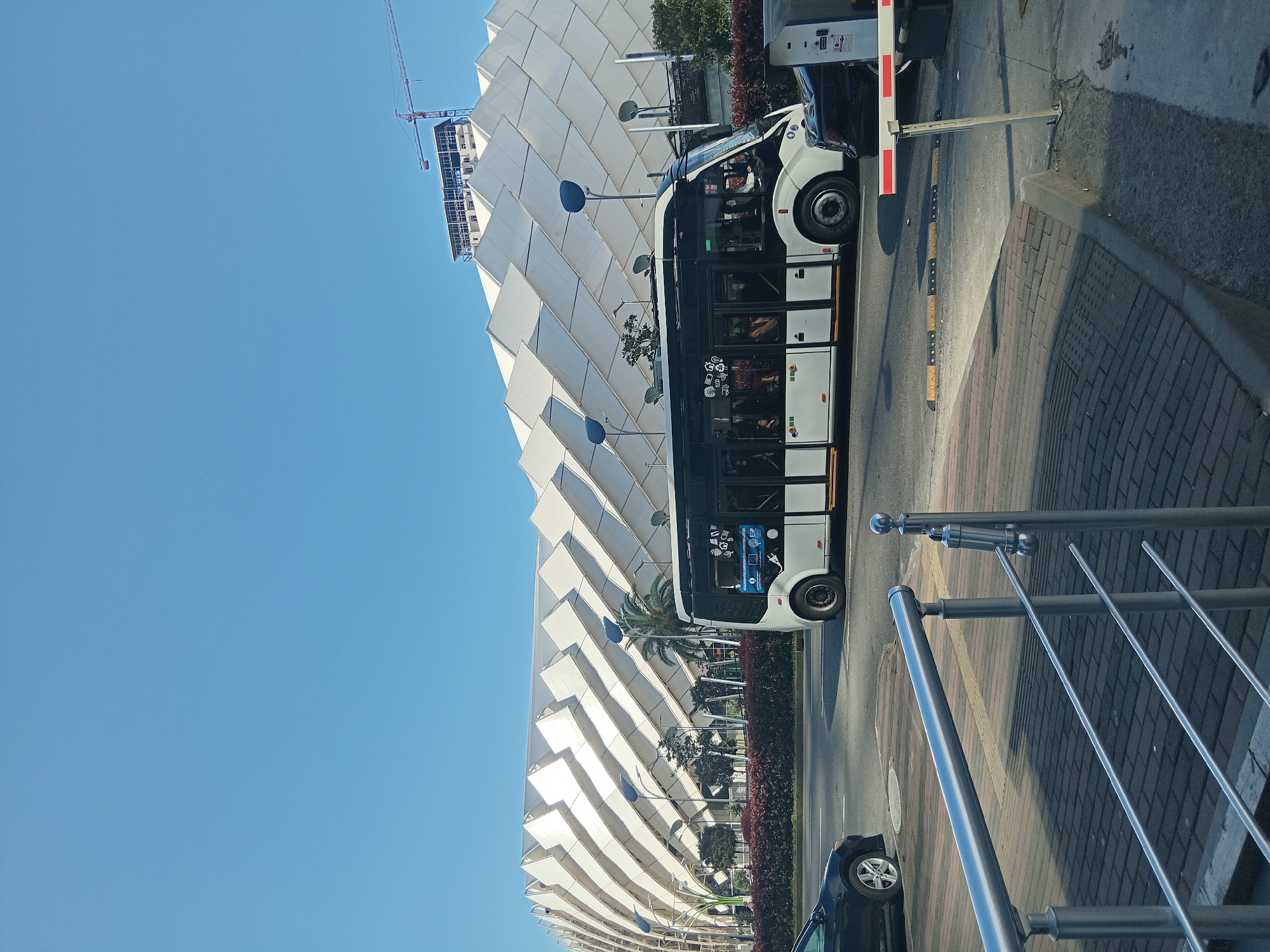 The image shows a white city bus stopped on a street in Batumi, Georgia, next to a modern architectural building. The building has a facade of stacked, white triangular shapes. A construction crane is visible above the building. A black car is parked behind the bus. A metal railing is on the right side of the frame, along with a red-brick sidewalk. The sky is clear and blue. The bus appears to be electric, based on the charging port graphic on the side. People are visible inside the bus.