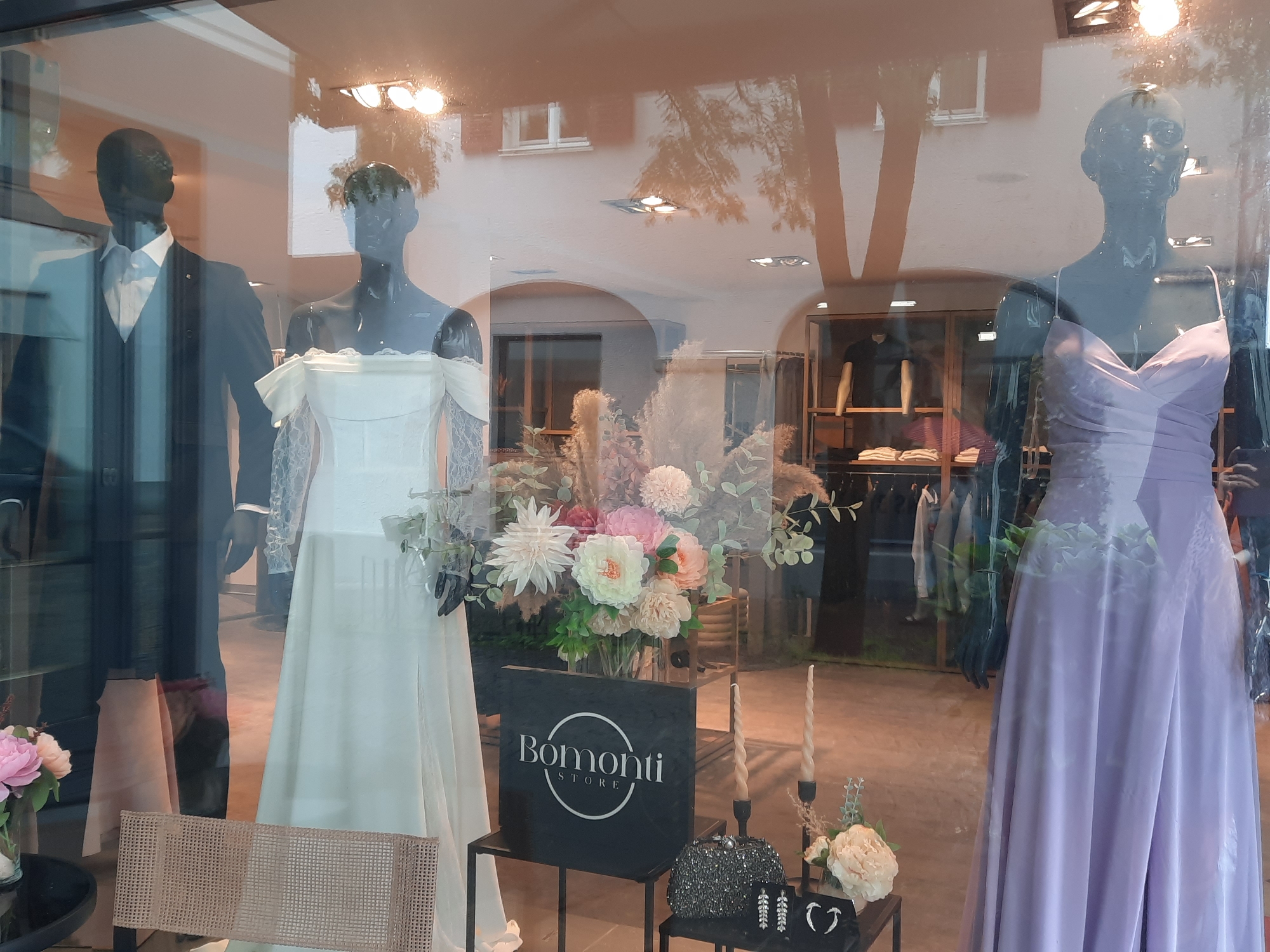 This image shows a store window display for "Bomonti Store" in Rosenheim, Germany. The display features three mannequins. The mannequin on the left is wearing a dark suit with a white shirt and bow tie. The middle mannequin is dressed in a white off-the-shoulder wedding gown with lace sleeves. The mannequin on the right wears a long, flowing lilac-colored dress.
In front of the mannequins, there is a black display board with the "Bomonti Store" logo. Beside it, a vase of artificial flowers in various shades of pink, cream, and white is arranged with dried grasses. To the right, there are two tall, twisted candles in black candle holders, and a small table displays a sparkly clutch purse, dangling earrings, and a heart-shaped jewelry piece. A woven chair is visible in the lower left corner.
The store's interior is visible through the window, showing clothing racks with garments. Reflections of buildings, trees, and lighting fixtures from across the street are prominent on the glass surface, obscuring some of the interior details. The overall impression is of an upscale boutique, likely specializing in formal wear or bridal attire.