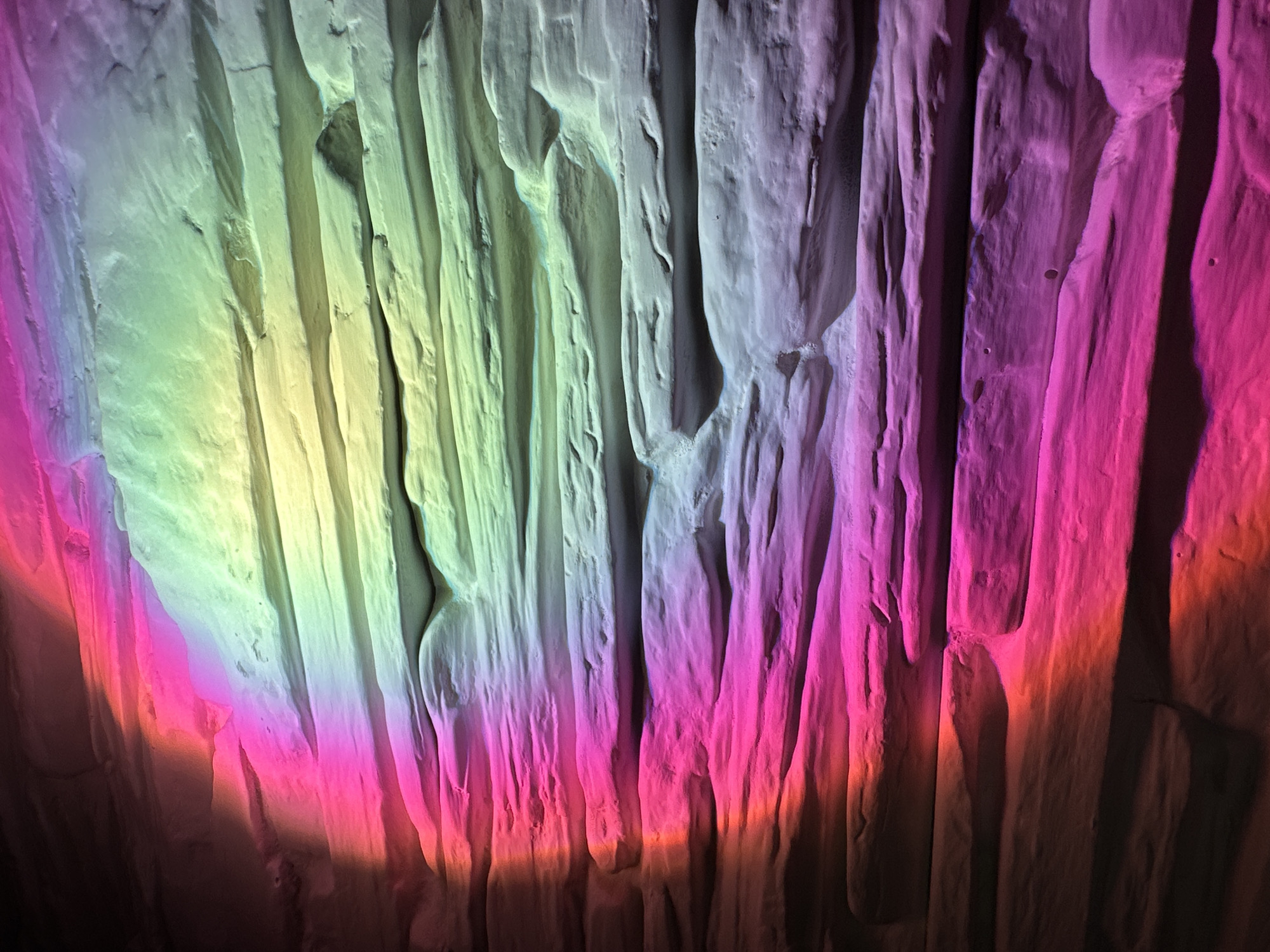 The image captures a close-up of a highly textured, vertically grooved surface, resembling a rock wall, cave formation, or abstract sculpture. The surface is dramatically illuminated by a spectrum of artificial, multi-colored lights.
From the left, the surface is bathed in bright yellow and lime green light, transitioning horizontally across to cooler blue-white tones in the center. Towards the right, the lighting shifts to vibrant magenta and deep pinks, with hints of orange and red appearing at the bottom and far right edges. This colored illumination highlights the rugged, uneven contours of the vertical ridges and channels, creating strong shadows that emphasize the three-dimensional depth of the texture. The surface itself appears solid and irregular, with various bumps, crevices, and smoother planes.
There are no discernible objects, people, or activities taking place within the frame. The background is dark, focusing all attention on the illuminated textured wall. The lighting suggests an interior setting, possibly a cave or an art installation designed to showcase the interplay of light and form. No text is visible in the image.