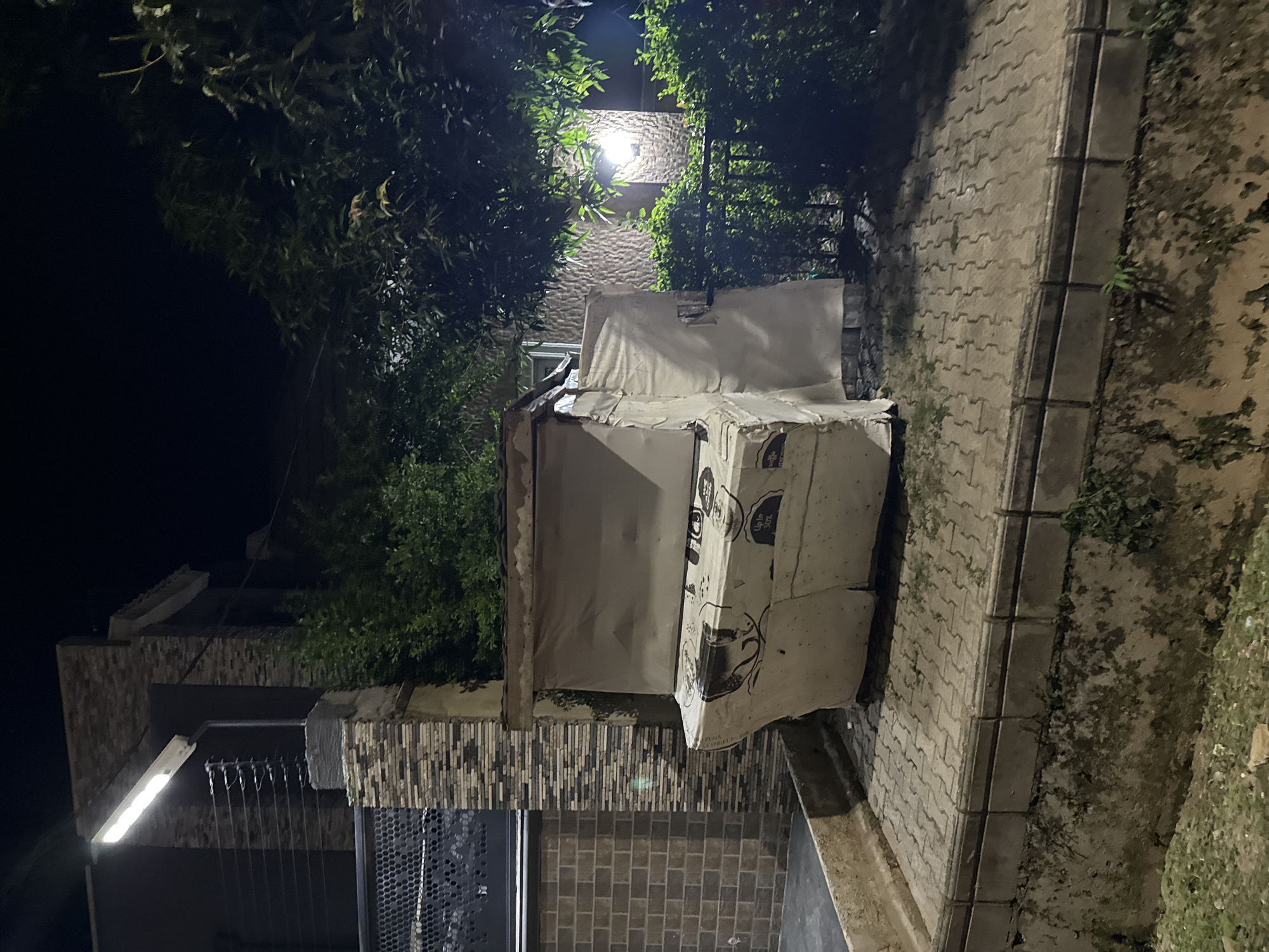 In Abuja, Nigeria, an upright piano, draped in white plastic sheeting, sits on a paved area at night. The piano is heavily wrapped, obscuring most of its details, but some text and imagery are visible on the lower section. Behind the piano, a lush green hedge and trees are illuminated by a warm light emanating from a wall-mounted lamp on a building. The building itself appears to be constructed with brick and stone. The paved area is bordered by a concrete ledge, below which is a vegetated embankment. The scene is dimly lit, with shadows cast by the surrounding foliage and building elements.