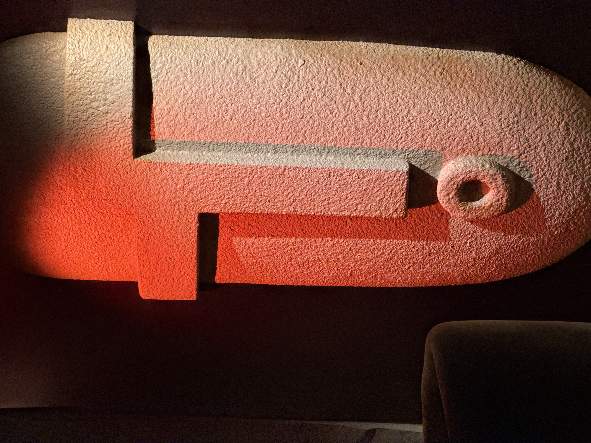 A close-up interior shot shows a textured, abstract, geometric shape on a wall. The shape is primarily light tan, with a pronounced pinkish-red hue from a light source emanating from the left side of the frame. The shape consists of rectangular and curved sections, and a circular feature on the right, all appearing to be made of the same textured material. The lower portion of the image is dominated by a dark, indistinct background, partially illuminated by the pinkish-red light. A portion of a dark-colored object, possibly a chair, is visible in the lower right corner. The environment is dimly lit.
The Moscow, Russia location is not stated in the image.