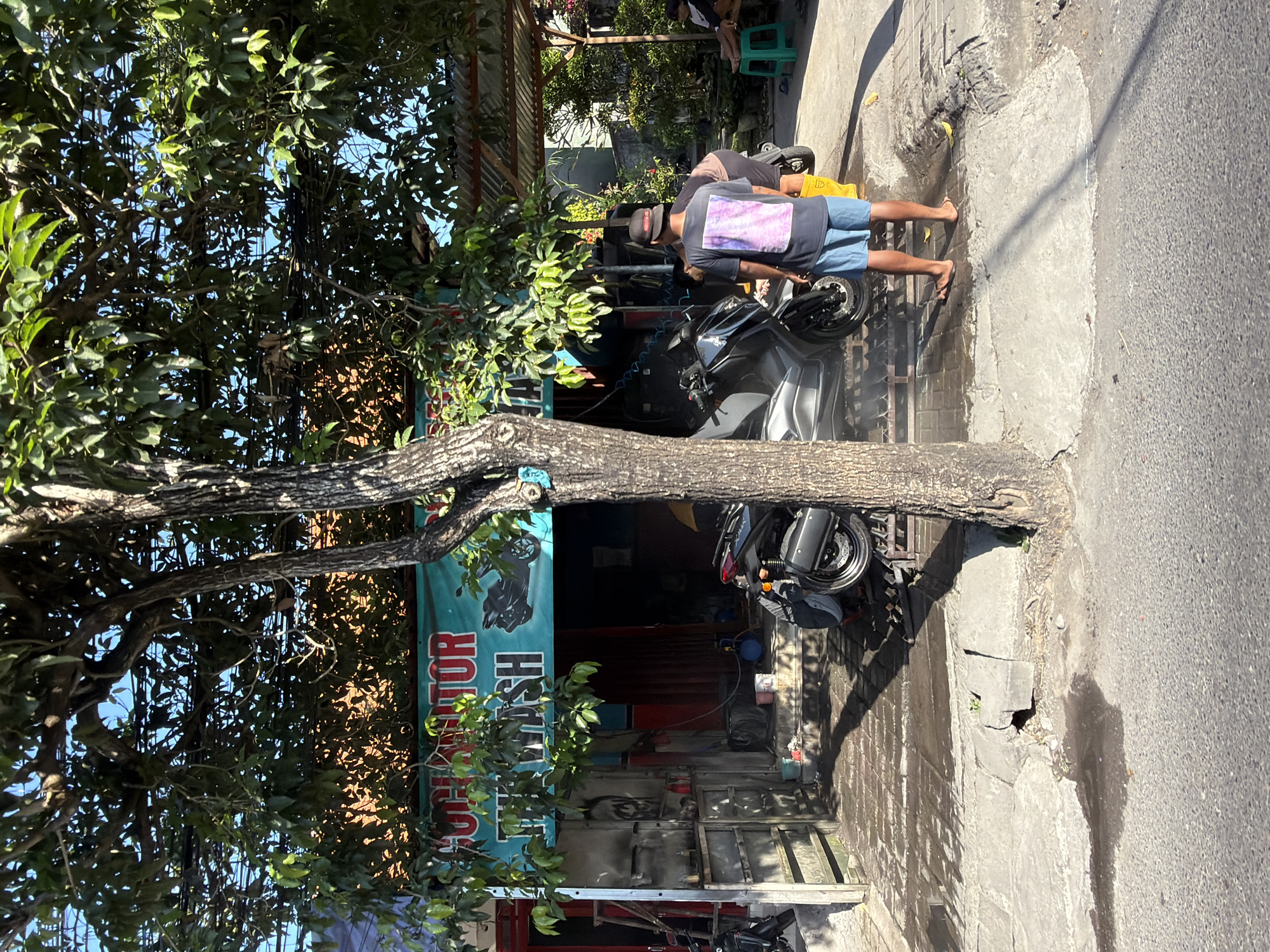 In North Kuta, Indonesia, a busy street scene unfolds outside a motorcycle wash establishment. A large tree with lush green foliage partially shades the area. A banner above the entrance reads "CUCI MOTOR," indicating "Motorcycle Wash." Inside the establishment, a dark-colored scooter is parked. A man wearing a black cap, a t-shirt with a pink and purple graphic, blue shorts, and yellow flip-flops stands near the scooter, appearing to be washing it. Another person, whose upper body is visible, is also attending to the motorcycle. To the right, a small child sits on a green stool. The ground is wet, suggesting recent or ongoing washing activities, with puddles reflecting the sunlight. The overall environment appears to be a typical street-side business in a tropical setting, with a mix of commercial and residential elements visible in the background.