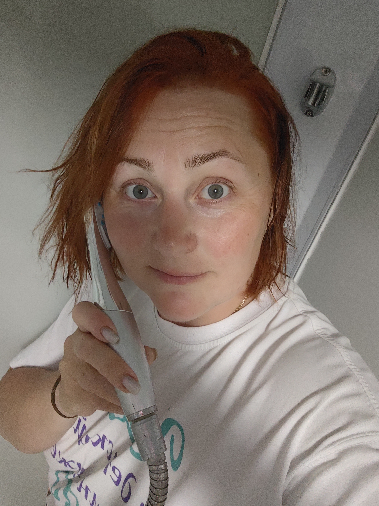 A woman with reddish-orange hair is holding a shower head to her left ear, as if using it as a phone. She is wearing a white T-shirt with a faint design. The background appears to be a clean, white interior, potentially a bathroom or shower stall. A metal fixture is visible in the upper right corner of the frame. The overall lighting is bright and diffused. Фоки, Russia, cannot be determined from the image.