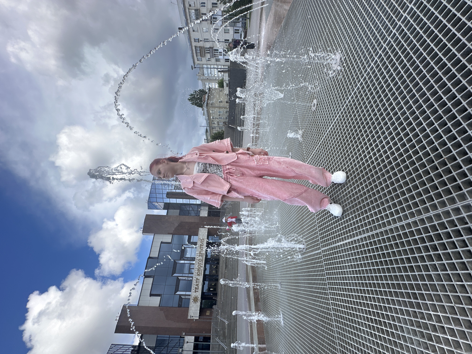 A young woman in a pink suit is standing in a fountain in front of a building with the Cyrillic text “ТЕАТР ЮНОГО ЗРИТЕЛЯ”, which translates to “Theatre of the Young Spectator”. The location is identified as Saratov, Russia. The woman is positioned amidst streams of water shooting upwards from the fountain's surface. The ground around the fountain consists of a grid-like metal surface. The sky behind the building is a mix of blue and cloudy skies. A taller building is visible in the background.