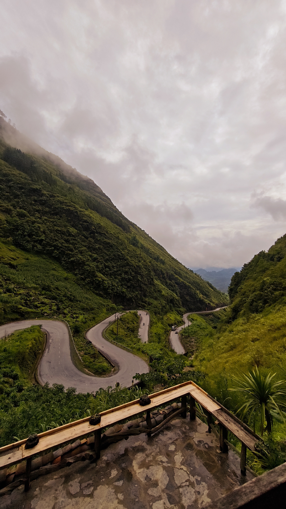 A high-angle shot shows a winding road in Phố Bảng Commune, Vietnam, set against a backdrop of lush, green mountains and a cloudy sky. The road snakes through the valley, with a couple of vehicles seen navigating the turns. In the foreground, a wooden structure, possibly a viewing platform, offers a vantage point to observe the landscape. The platform has a weathered appearance, with evidence of moisture. The overall atmosphere is one of rural tranquility, punctuated by the road's infrastructure.