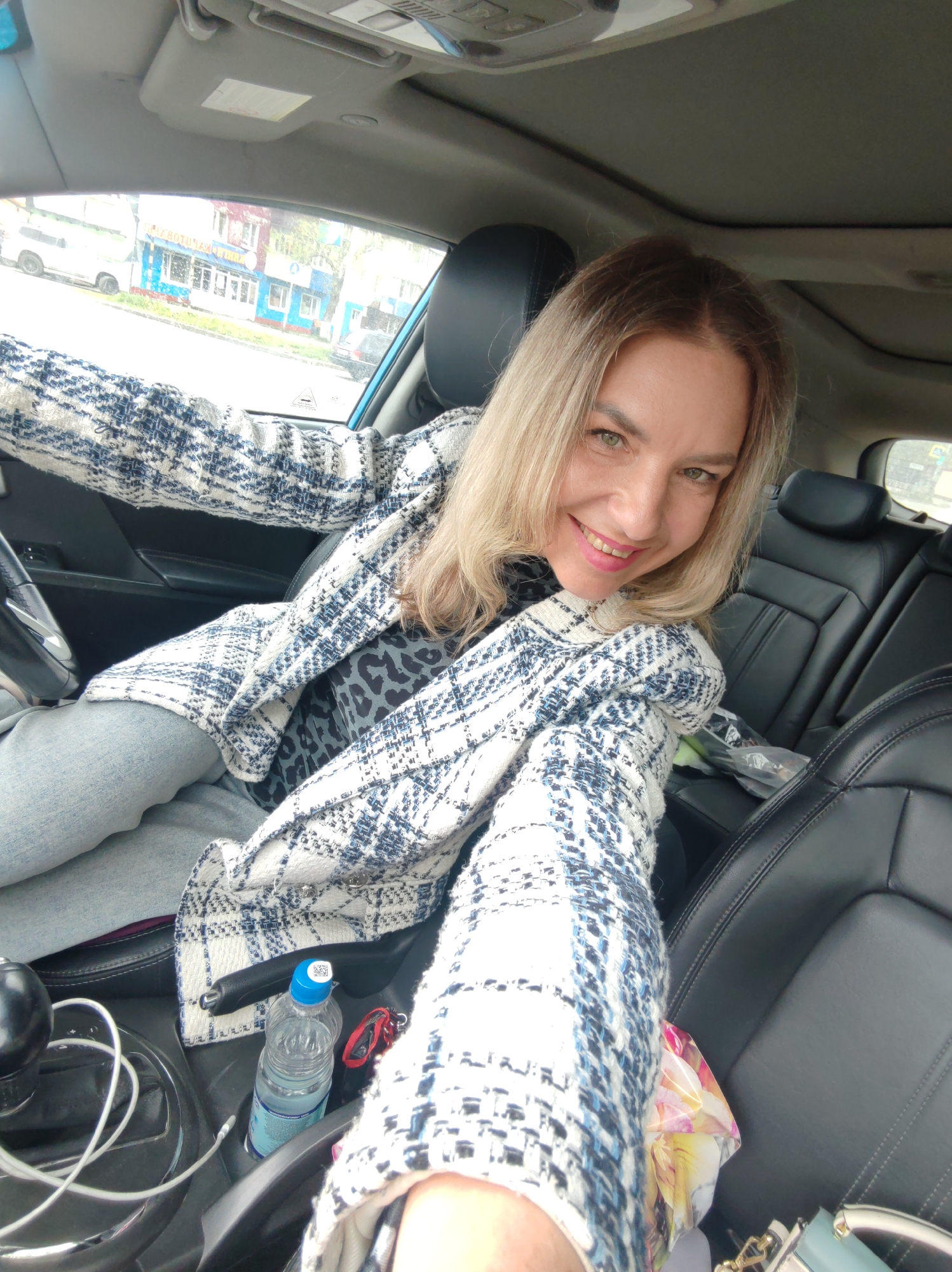 A fair-skinned woman with blonde hair is sitting in the driver's seat of a car and taking a selfie. She is wearing a white and blue patterned jacket and a leopard print shirt. The car's interior is visible, with black leather seats and a steering wheel. Outside the car window, a street scene with buildings and vehicles can be seen. The image appears to have been taken in Petropavlovsk-Kamchatsky, Russia, as indicated by the presence of a sign with Cyrillic text.