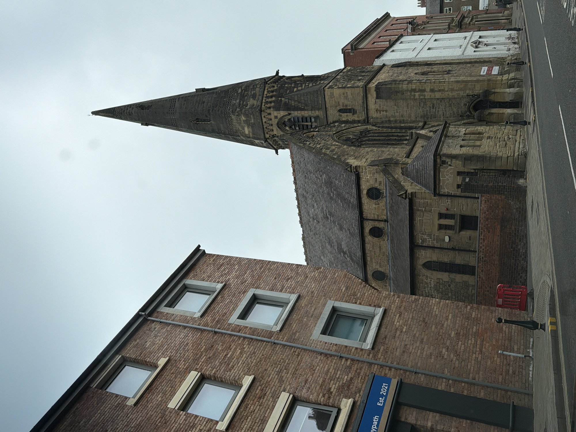 The image shows a view of the city of Durham, United Kingdom. In the center stands St. Nicholas Church, distinguished by its tall, conical spire constructed from weathered stone. The church is adjoined by buildings of varying architectural styles, including a red brick structure with modern, square windows and a building with red and white brick patterns. A black street lamp stands along the edge of a paved path and a road. The sky is overcast, giving the scene a muted, gray tone. A sign on the modern brick building says "Mypass Est. 2021" with a blue background.