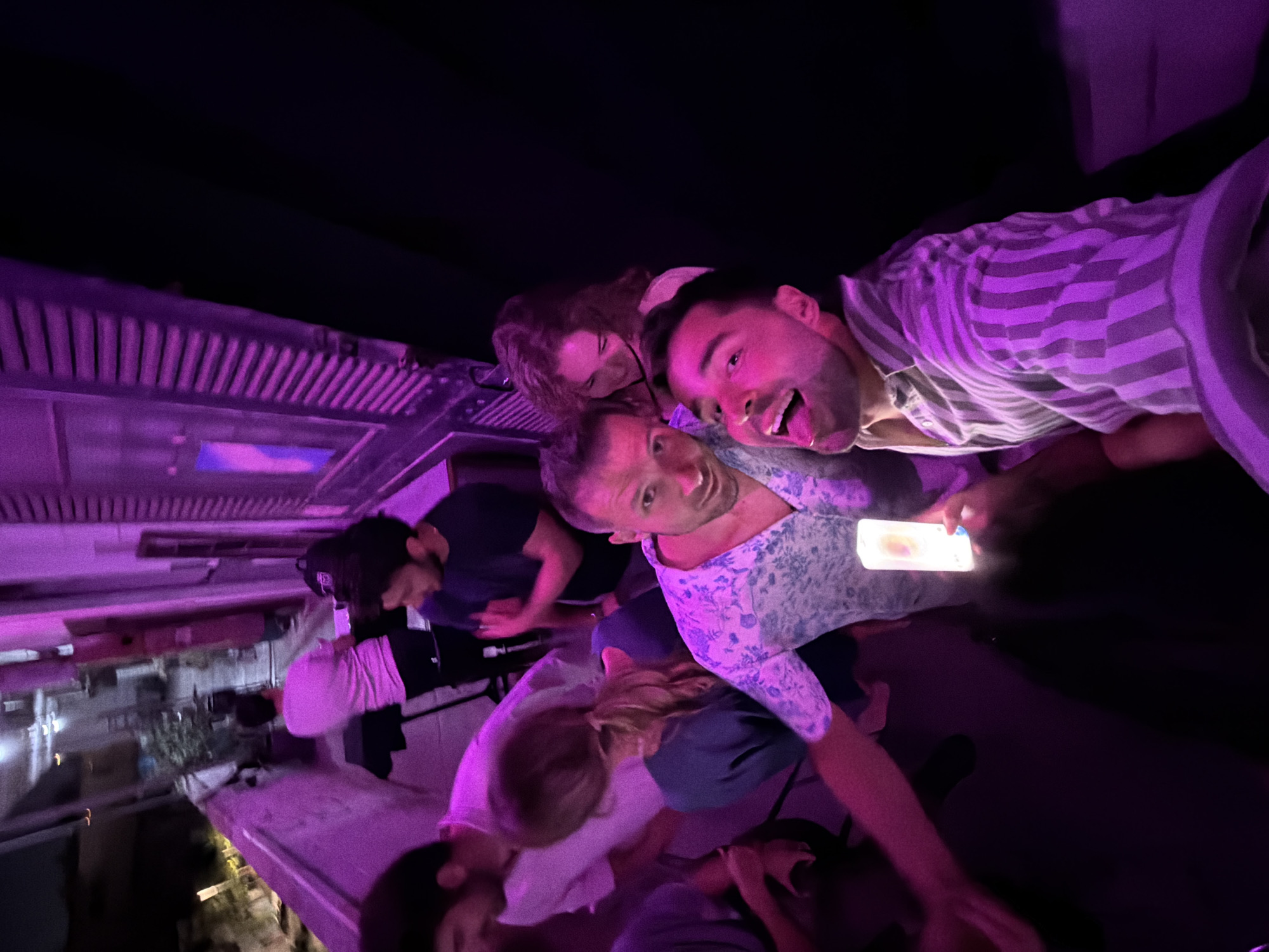 This detailed description is based on the provided image and crops, with the city identified as Tel-Aviv, Israel, as requested.
The image depicts a nighttime outdoor social gathering on what appears to be a balcony or rooftop in Tel-Aviv, Israel. The scene is illuminated predominantly by artificial purple light, casting a distinct hue over the subjects and immediate surroundings.
In the immediate foreground, a man with short dark hair is seen from the chest up, wearing a vertical striped shirt (white and grey/purple). He grins widely at the camera with his tongue extended. He holds a glowing smartphone in his right hand, angled upwards. To his left, another man with short dark hair, wearing a white shirt with a light blue floral pattern, also looks towards the camera with a slight smile.
Behind these two men, slightly to the right, a woman with curly brown hair smiles and looks downwards. Another woman is partially visible behind her to the far right. Further to the left in the mid-ground, a woman with blonde hair tied in a ponytail is seen from the side, wearing a dark blue top and looking downwards. Adjacent to her, a man in a light pink t-shirt faces forward, while another man in a dark t-shirt is visible from the side, looking left with his hands clasped or gesturing.
In the background, on the far left, a person in a light-colored t-shirt is positioned near a low wall or railing, seemingly operating audio equipment on a stand or table, which also holds a dark backpack. The outdoor setting includes a distant view of blurred city lights and buildings against a dark sky, visible beyond the railing on the left. To the right, part of a building facade with vertical shutters is visible. The pervasive purple lighting suggests a festive or entertainment event.