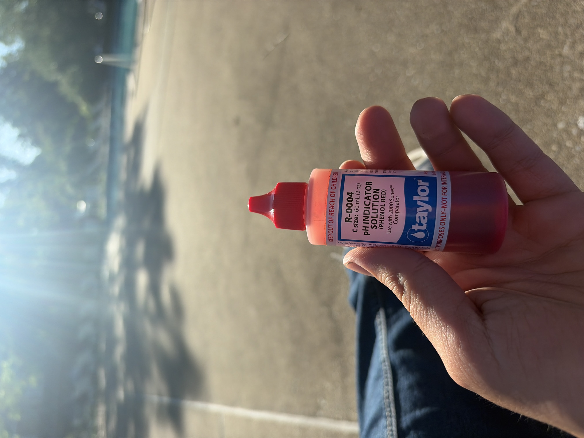 A person's hand holds a Taylor R-0004 pH Indicator Solution (Phenol Red) in an outdoor setting. The solution comes in a red plastic bottle with a red cap, bearing the Taylor logo and product information, including the volume of 60 mL (2 oz). The label also states "KEEP OUT OF REACH OF CHILDREN" and "TEST PURPOSES ONLY - NOT FOR INTERNAL USE." The person is wearing blue jeans. The background consists of an out-of-focus area with hints of greenery, suggesting trees or vegetation, and a lighter-colored surface possibly indicating a sidewalk or pavement. The image suggests the photo was taken outdoors on a sunny day, given the strong sunlight and shadows. The label indicates Taylor Water Technologies LLC at 31 Lovelon Circle. Edison, United States of America.