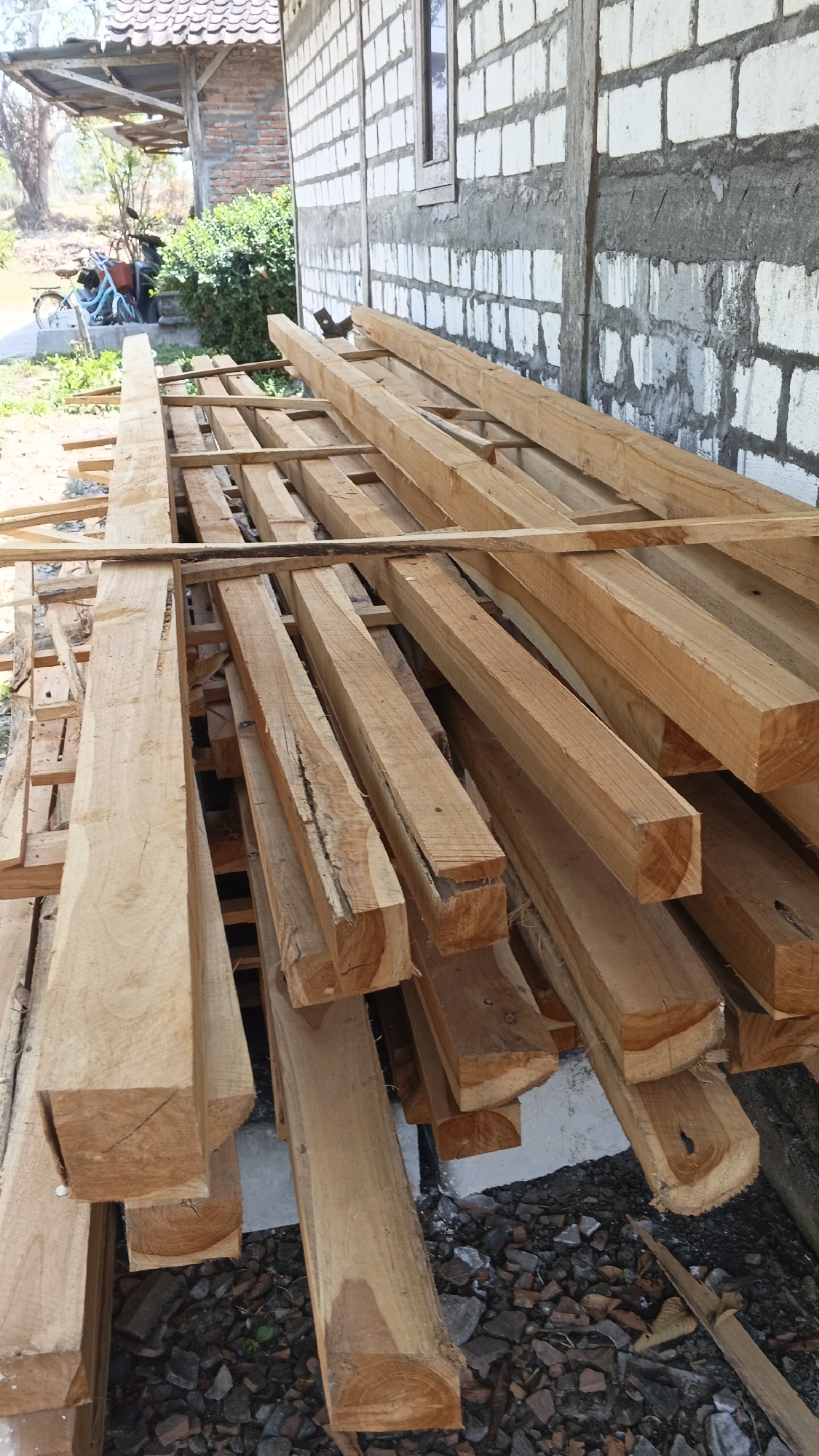 A vertical, medium shot shows a pile of rough-hewn lumber stacked in front of a building in the village of Soko, Indonesia. The lumber is light brown, with visible grain and some cracks. The building behind the lumber has white brick walls with gray mortar and a small window. To the left of the building, a covered porch area is visible, with a blue bicycle and a motorcycle parked underneath. The ground is covered with dirt and small stones. The lighting suggests it is daytime.