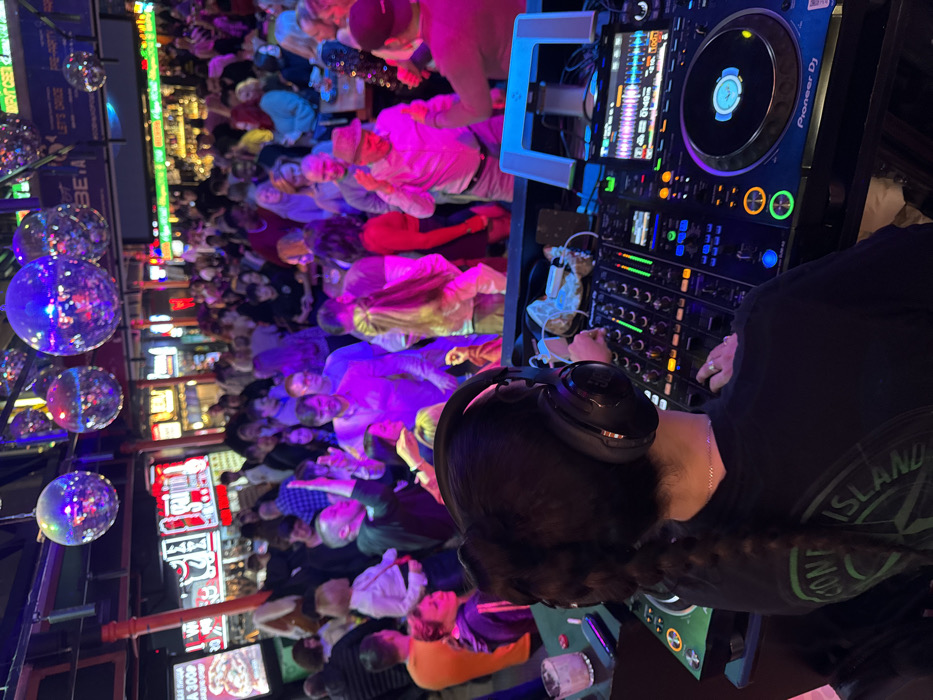 The image captures a crowded nightclub or bar scene in Moscow, Russia, evidenced by signage displaying "ПИЦЦА 300P" (Pizza 300 Rubles).
In the foreground, a person, identified as a DJ and viewed from behind, operates a professional DJ setup including a Pioneer DJ mixer and at least one Pioneer DJ media player. The DJ wears black JBL over-ear headphones and a dark t-shirt with a partially visible "STONE ISLAND" logo on the back. Their dark hair is styled in a long braid. A clear glass containing a beverage and ice, along with a mobile phone, rests on the DJ console to the left.
The venue is dimly lit but intensely illuminated by various colorful LED and neon lights, predominantly in shades of pink, purple, green, and red. Multiple disco balls are suspended from the ceiling, reflecting these lights across the space. The midground and background are densely populated with a diverse crowd of patrons engaged in dancing and socializing. Visible individuals include a man in a pink shirt and fedora on the right, a man in a light purple shirt near the center, and a person holding a small child on the left. The background also reveals additional bar areas and illuminated signage, some in English, such as "DRINK CREW" and "LET'S DANCE."