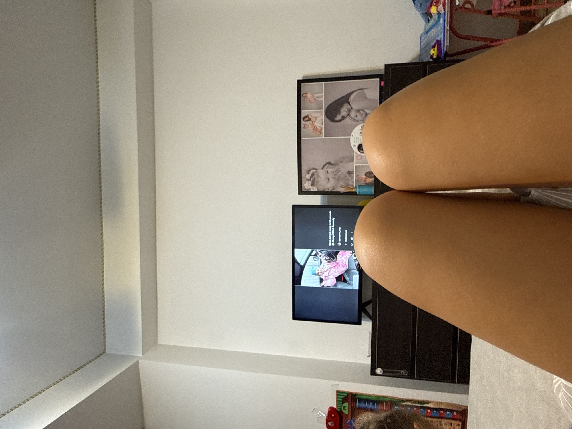 The image depicts a bedroom setting, likely residential, with a focus on a dresser and two screens. On the left screen, a person in a pink outfit is visible inside a vehicle. On the right, a framed collage of photos is displayed, featuring images of a baby and a woman. In the foreground, two legs are positioned, suggesting the viewer is lying down. The overall impression is one of a casual, indoor environment. The location of Chișinău, Moldova, is not discernible from the visual elements present in the image.