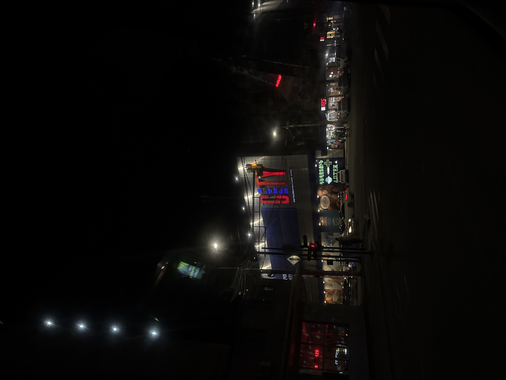 The image depicts a street scene at night in Yerevan, Armenia. A large billboard dominates the view, brightly lit with Cyrillic script, possibly advertising a store or business. The billboard is mounted on a building with red neon signs and illuminated displays. Streetlights illuminate the scene. Further down the street, a KFC restaurant is visible. There are a few pedestrians visible on the right side of the image. The image was most likely taken from a moving car.