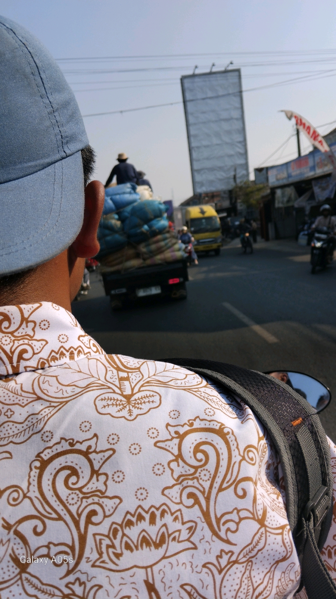 The image captures a perspective from someone riding on a motorcycle in Sumedang, Indonesia. The foreground is dominated by the back of the rider's head, wearing a light blue baseball cap, and their white shirt adorned with a detailed brown paisley and floral pattern. A grey backpack strap is visible crossing the shoulder. In the middle ground, a black pickup truck is seen carrying a large load of sacks, possibly agricultural produce, wrapped in blue tarpaulin. A person wearing a dark blue shirt and a hat is perched on top of the load. Further down the road, a yellow truck is traveling in the same direction, and several other motorcycles are also visible on the street, indicating moderate traffic. The background shows buildings, overhead power lines, and a tall, rectangular billboard structure. The sky is clear and blue, suggesting a sunny day. The scene appears to be on a paved road in an urban or semi-urban area.