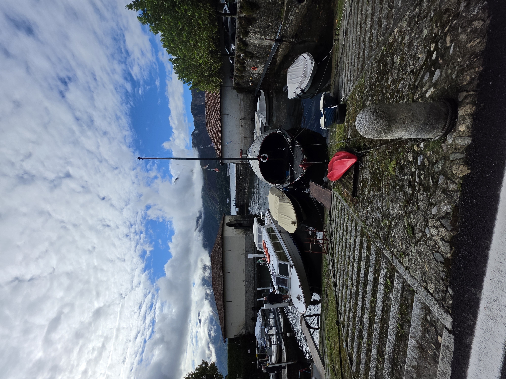 The image depicts a harbor in San Giovanni, Italy, under a cloudy sky with patches of blue. Several boats are docked, including a large, dark boat with a covered cabin and a white passenger boat with orange life preservers. A man in a dark shirt stands on the passenger boat, facing away from the camera. In the background, there are stone buildings with terracotta roofs and lush green trees. Mountains are visible in the distance. The foreground features a stone staircase leading down to the water. A red kayak lies on the stone embankment.