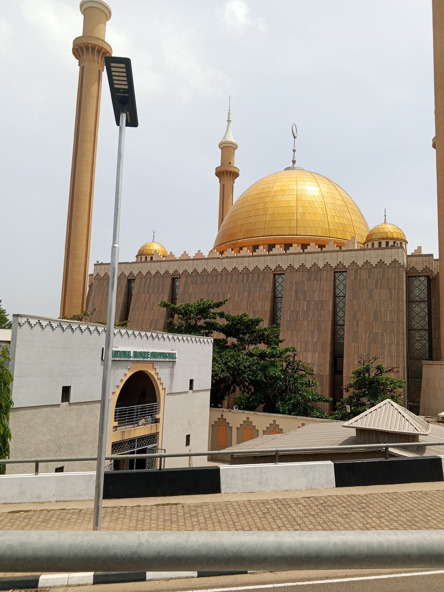The image depicts the National Mosque of Nigeria, located in Abuja. The mosque features a large golden dome, surrounded by four minarets. The main structure of the mosque has a facade of brown panels. A building adjacent to the mosque features the signage "NATIONAL MOSQUE SHOPPING MALL." A modern street lamp is positioned in the foreground. The scene is captured during daylight hours with a slightly overcast sky. The foreground shows a street and barrier. No people are immediately visible in the image.