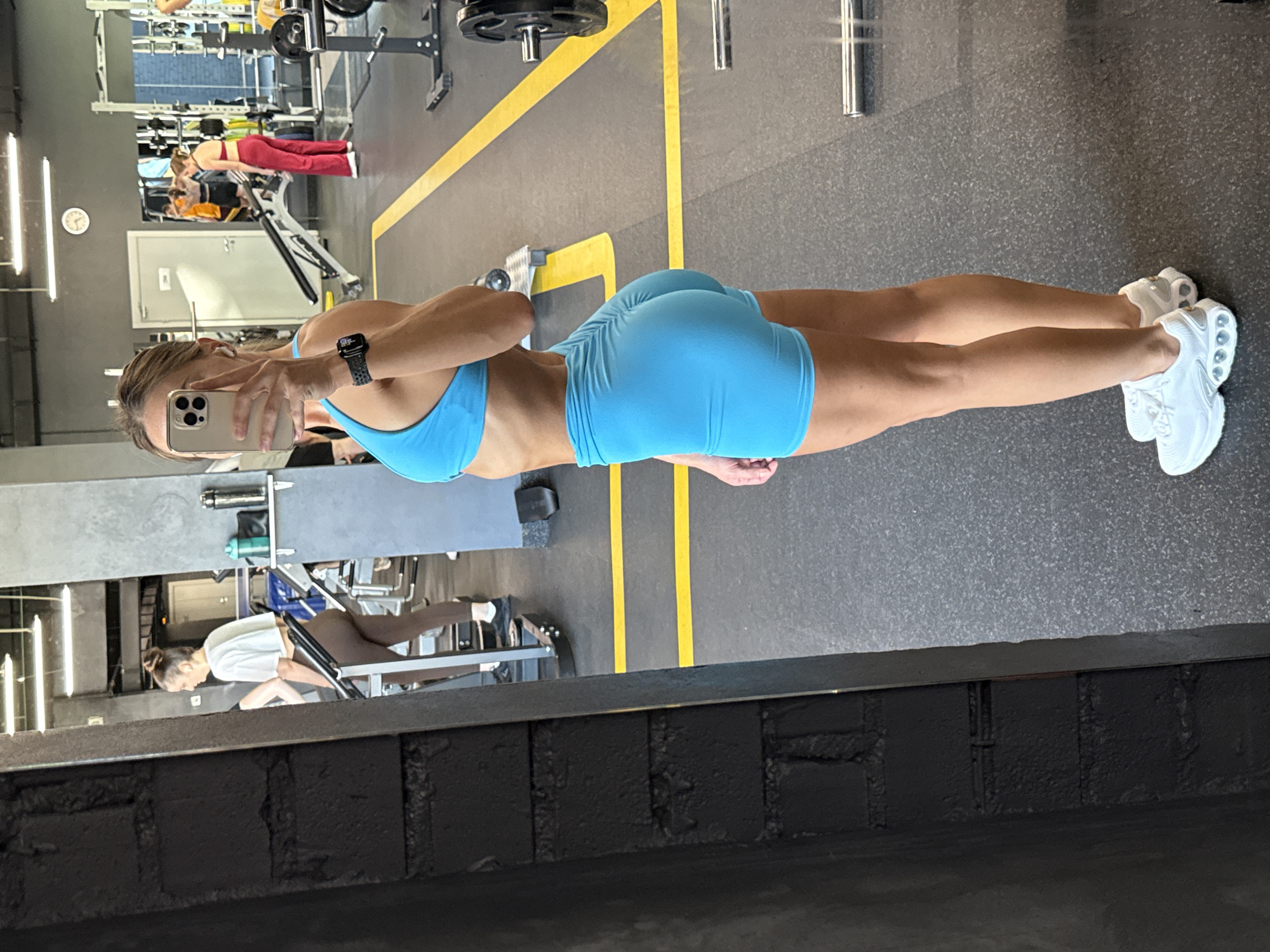 The image shows a woman in a gym. She is wearing a blue sports bra and blue shorts. She is holding a phone in her left hand and taking a selfie. Her right arm is bent at the elbow and resting on her hip. She is wearing white athletic shoes. In the background, there are gym machines and other people working out. The gym appears to be well-lit and equipped. The overall impression is of a fitness-focused environment. The reflection in the mirror suggests that the photo was taken at eye level. The woman's posture and expression do not convey any specific emotion.
