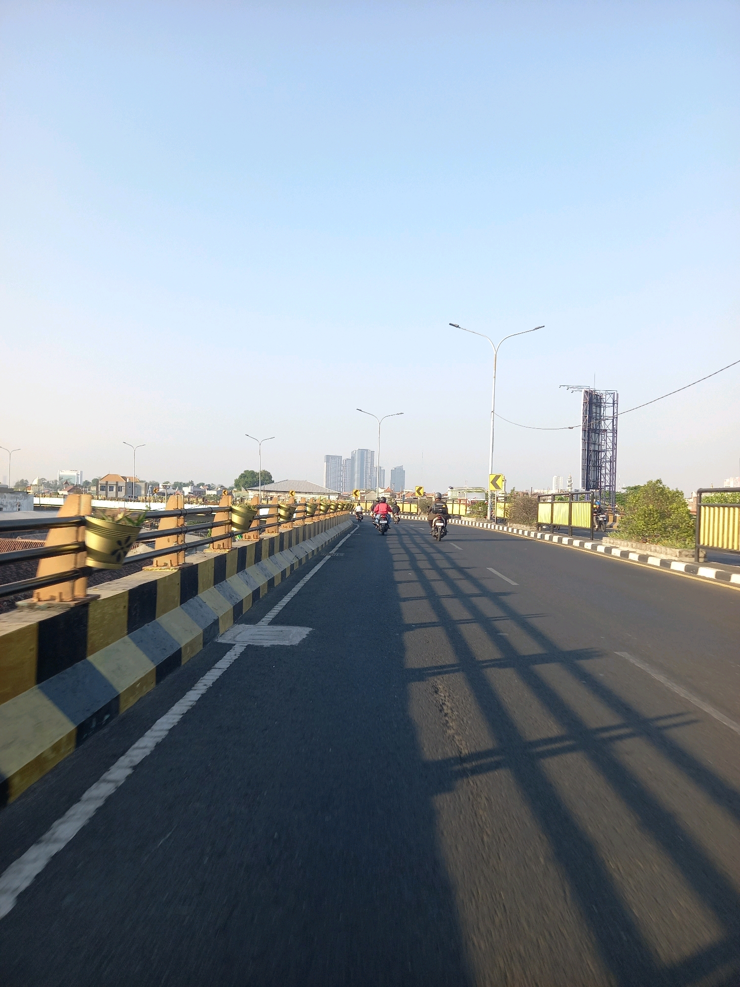 The image shows a street view in Surabaya, Indonesia, likely on a bridge, with a clear blue sky. The foreground features an asphalt road with a white line marking the lane. A safety barrier with yellow and black checkered patterns runs along the left side of the road, with decorative planters mounted on it. In the distance, several buildings, including some high-rise structures, are visible. The scene is illuminated by natural light, casting long shadows of unseen objects across the road. Several motorcycles, each with a rider, are moving along the road in the same direction. Streetlights are also present, lining the road.
