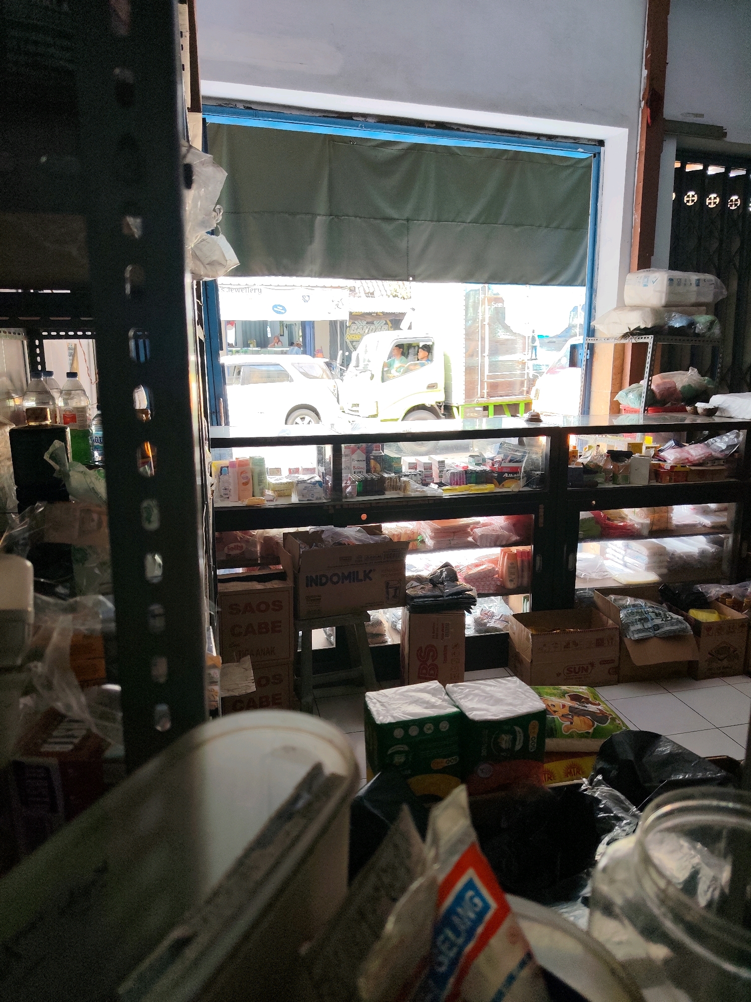 The image depicts the interior of a small store, likely a minimarket or grocery store, located in Dampit, Indonesia. The store appears to be cluttered with various goods stacked on shelves and on the floor. In the foreground, a metal shelving unit is visible on the left, filled with bottles of liquids and plastic-wrapped items. In the center, a glass display case is filled with packaged food items and toiletries. Boxes of products are also stacked on the floor, including a box labeled "INDOMILK" and another with "SAOS CABE" (chili sauce).
Through an open shutter or door in the background, a street scene is visible. A white truck with people inside is parked outside, and other vehicles and buildings can be seen. The overall impression is of a busy, functional retail space. The lighting suggests it is daytime.