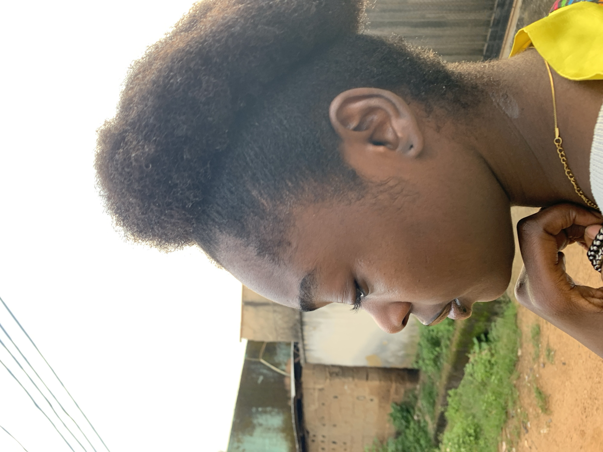 A profile shot captures a young Black woman from the nose upwards, looking towards the left. Her dark, curly hair is styled in a high bun. The background features a rustic, urban environment, likely within Rafin Par, Nigeria. Buildings constructed from brick and stucco are visible, some showing signs of age. Greenery, including grass and possibly a drainage ditch, lines the foreground, with a dirt path indicating a rural or unpaved road. The sky is bright and overcast, with power lines stretching across the upper portion of the frame. The woman is wearing a gold chain and a garment with a bright yellow collar. Her left hand is raised, with her fingers lightly touching her chin.