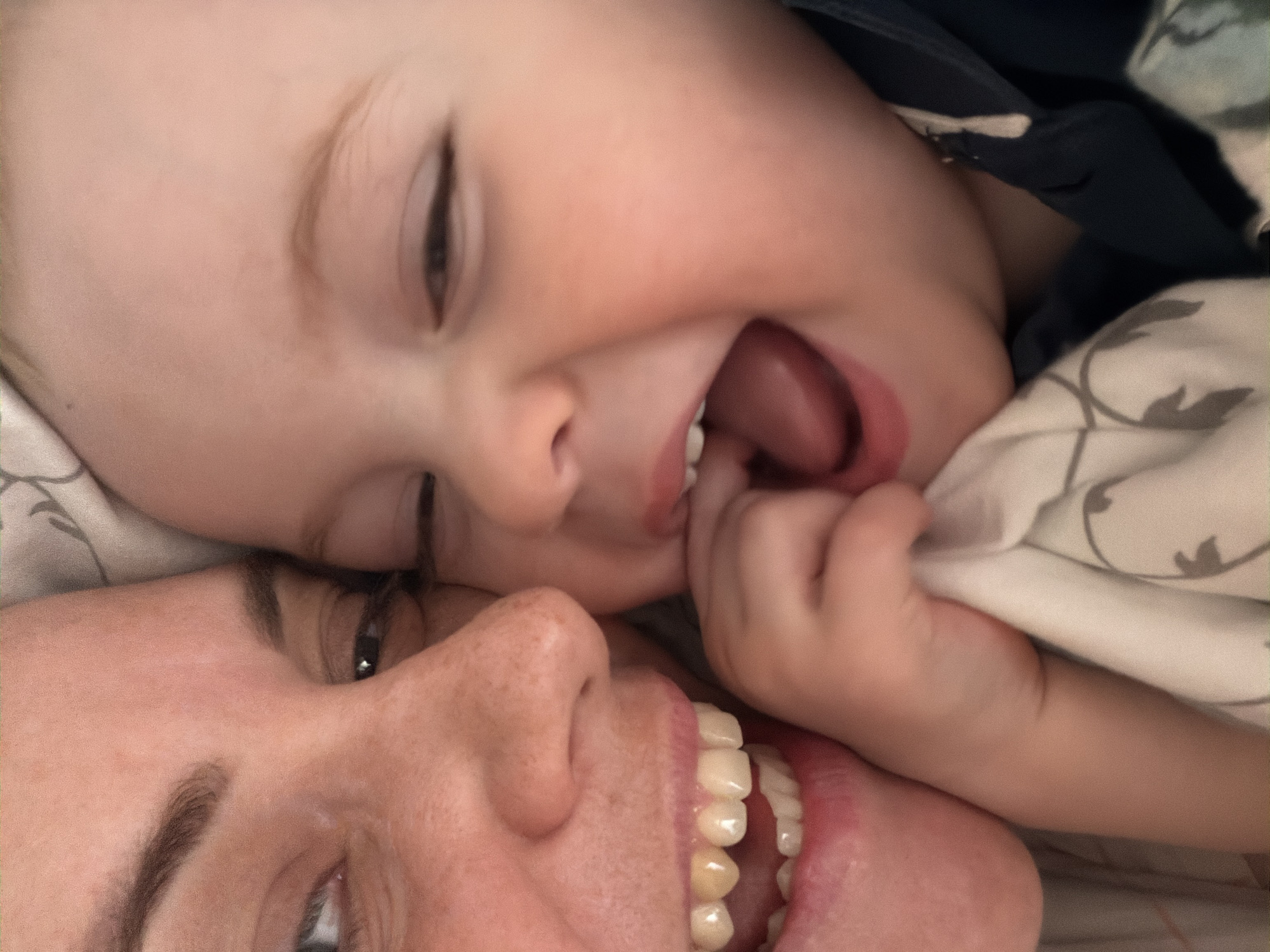A close-up, tilted image shows a woman and a child lying down, possibly in bed. The woman is facing the camera with her eyes partially closed and her mouth open, revealing her teeth. The child is lying above her, also with their eyes partially closed and mouth open, tongue visible. The child is holding a light-colored blanket with a pattern. The background appears to be bedding. The location is not discernible from the image and cannot be assumed to be Votkinsk, Russia.