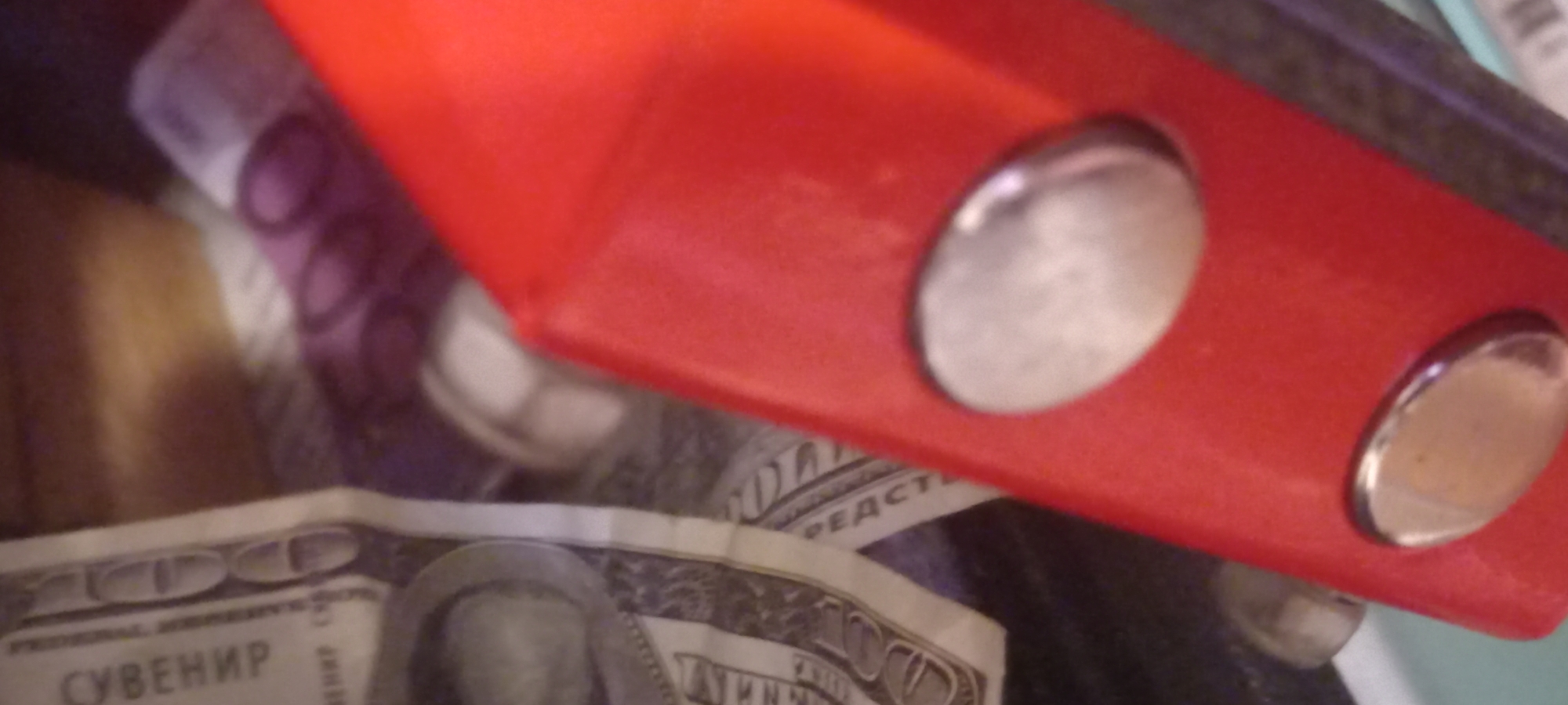 This is a close-up, slightly angled, low-resolution image of money and part of a red object. In the background, there are banknotes, including what appears to be a US $100 bill with text "СУВЕНИР" visible vertically. There are also hints of purple banknotes. The red object in the foreground is a thick, solid material with two oval, metallic or reflective buttons visible on its surface. The scene is indoors and appears to be in a cluttered environment. There is no visible indication of the city of Kryvyi Rih, Ukraine.