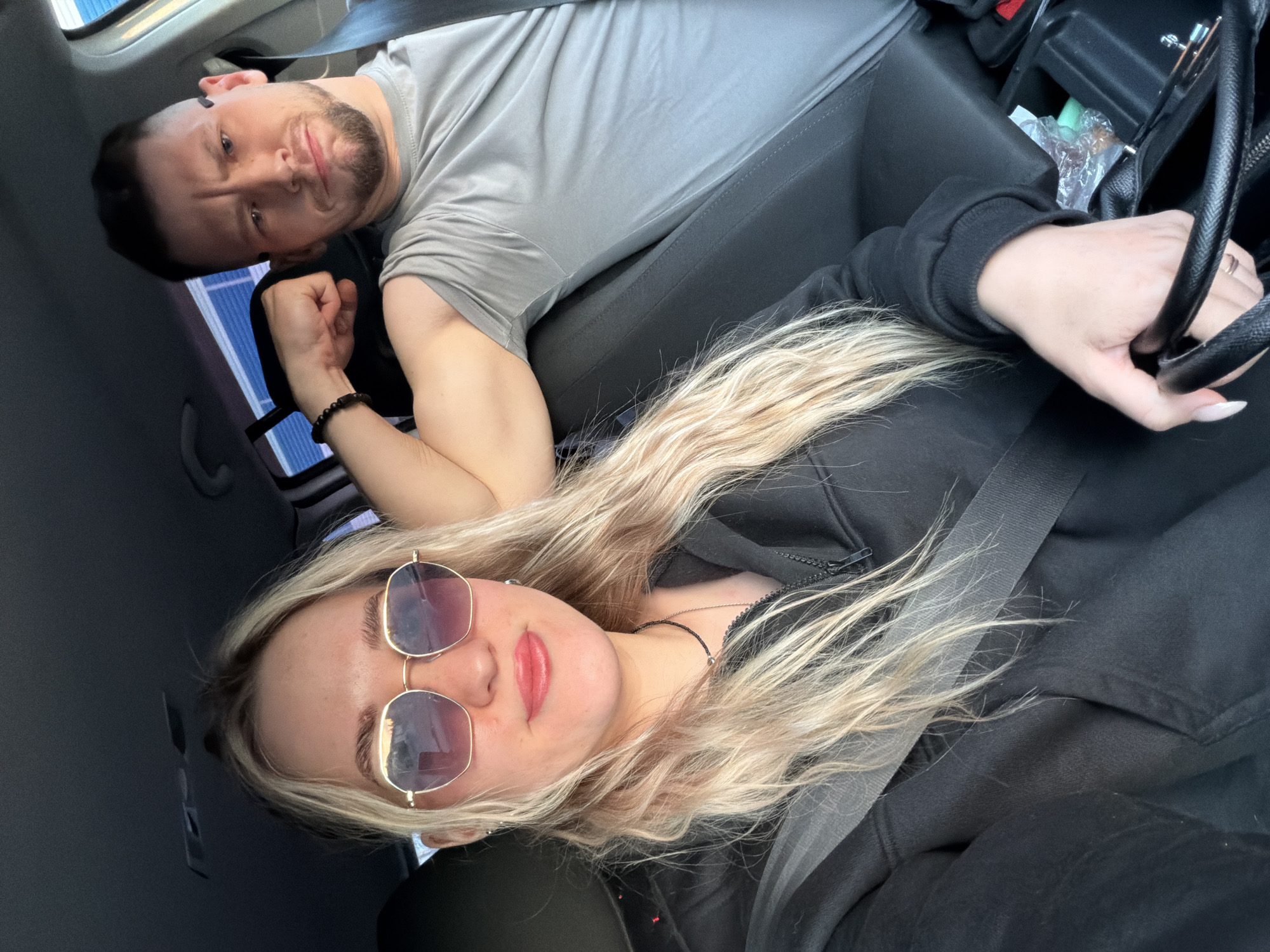 The image depicts two individuals inside a vehicle. A male with short, dark hair and a beard is wearing a gray shirt and a beaded bracelet on his left wrist. He is positioned in the background, closer to the window. In the foreground, a female with long, blonde hair and sunglasses is wearing a black hoodie. She is holding a black bag. Both are wearing seatbelts. The location appears to be inside a car, with a vehicle interior visible, including the dashboard and center console. The city and specific location are unknown.