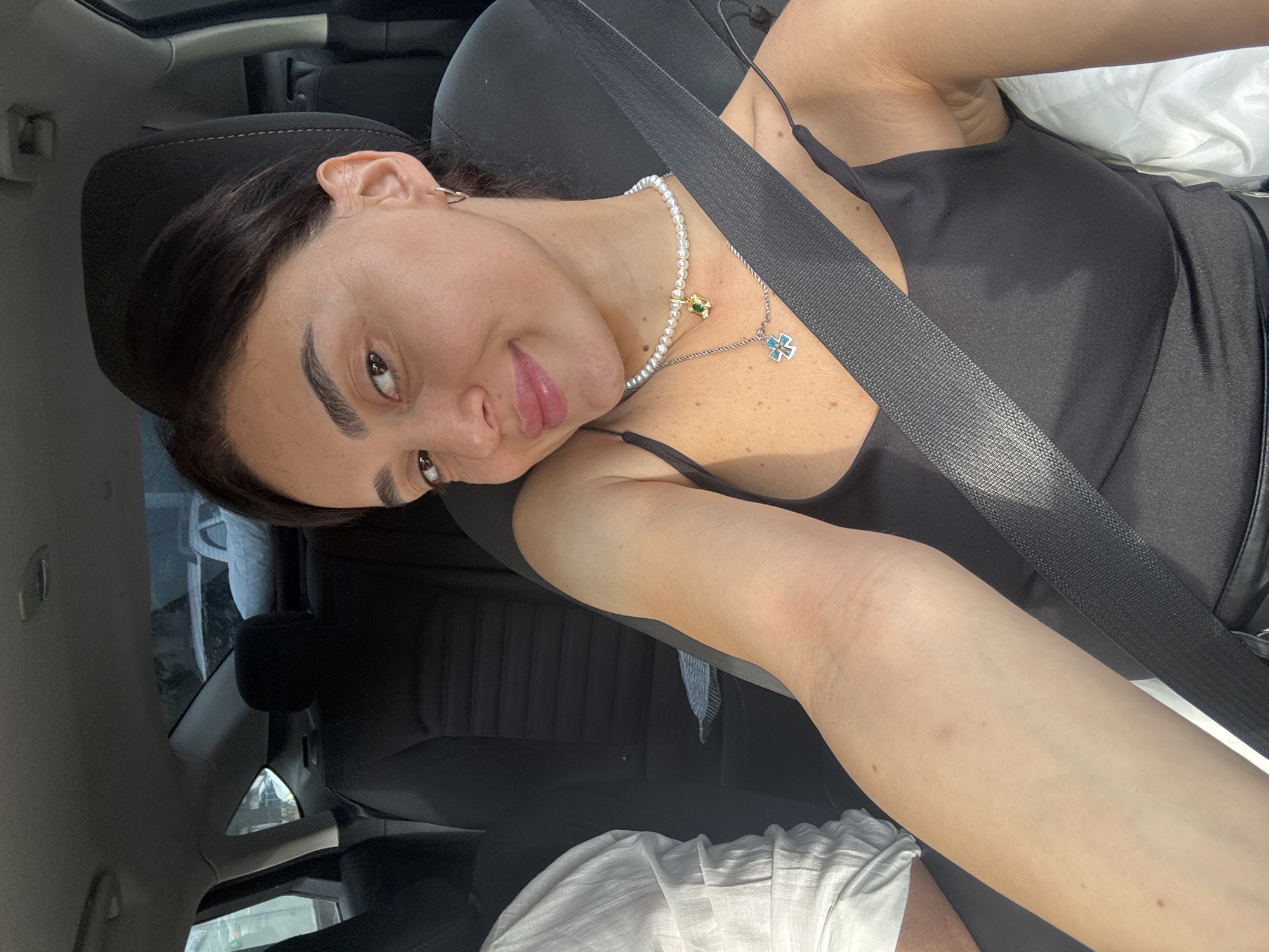 A young woman with dark hair and brown eyes is sitting in the passenger seat of a car. She is wearing a black tank top and a pearl necklace with two pendants. A seatbelt is fastened across her chest. The interior of the car is visible, including the black upholstery of the seats and the ceiling. Outside the car windows, trees and buildings can be seen, suggesting an urban environment. The lighting indicates it is daytime. The location is identified as Tbilisi, Georgia.
