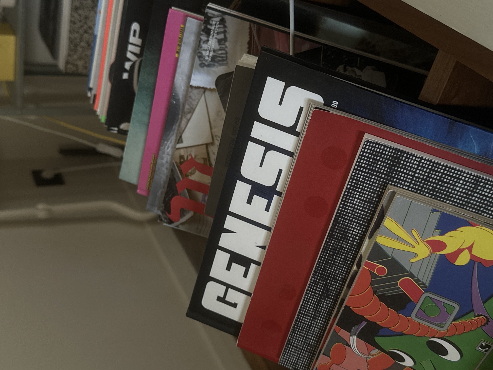 A close-up, canted shot shows a row of records in a wire record rack. The nearest record is a colorful, cartoonish illustration dominated by green, red, yellow, and blue hues. It sits behind a red record cover. Next to this is a record with the word "Genesis" written vertically in large, white letters on a black spine. Other records with varying colors and titles are visible behind it. The context for Saint-Ouen-sur-Seine, France, is absent from the provided information.