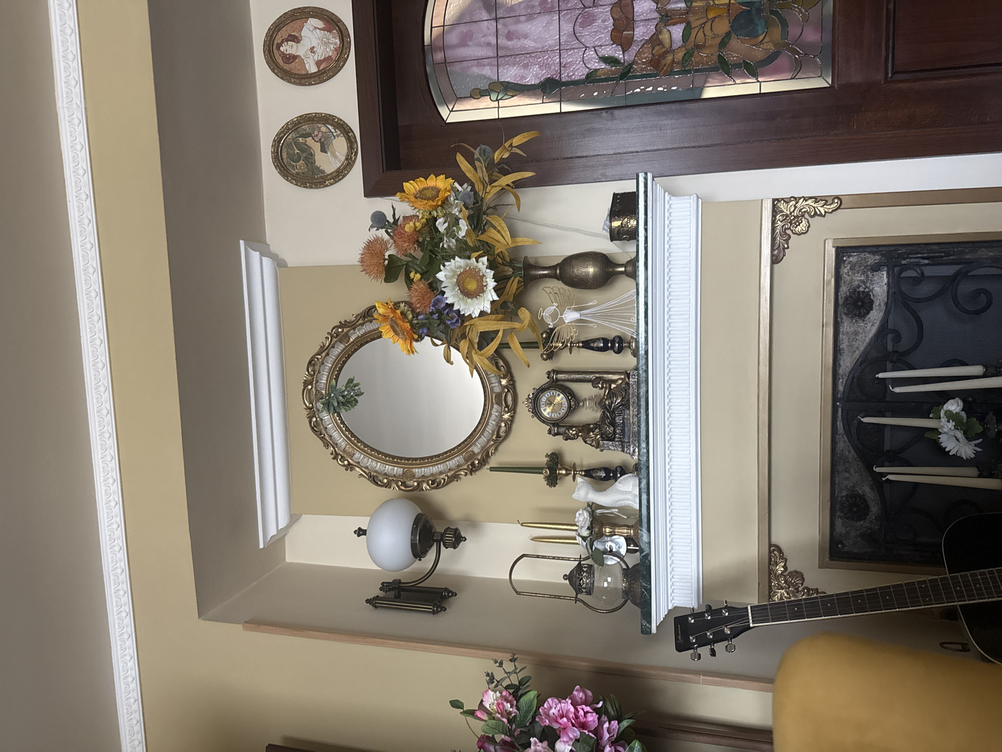 The image depicts an interior setting, likely a residential space in Trnava, Slovakia. A decorative display is arranged on a ledge or shelf, featuring an oval mirror with a gold frame, a floral arrangement with sunflowers and white flowers, several ornate candle holders with candles, and a vintage-style lamp with a white globe shade. Above the display, there are two round decorative plates with portraits on the wall, and a stained-glass window panel. To the right of the display, a guitar leans against the wall, partially obscuring a framed piece, possibly a painting or decorative panel with lit candles. The overall decor suggests a classical or vintage aesthetic.