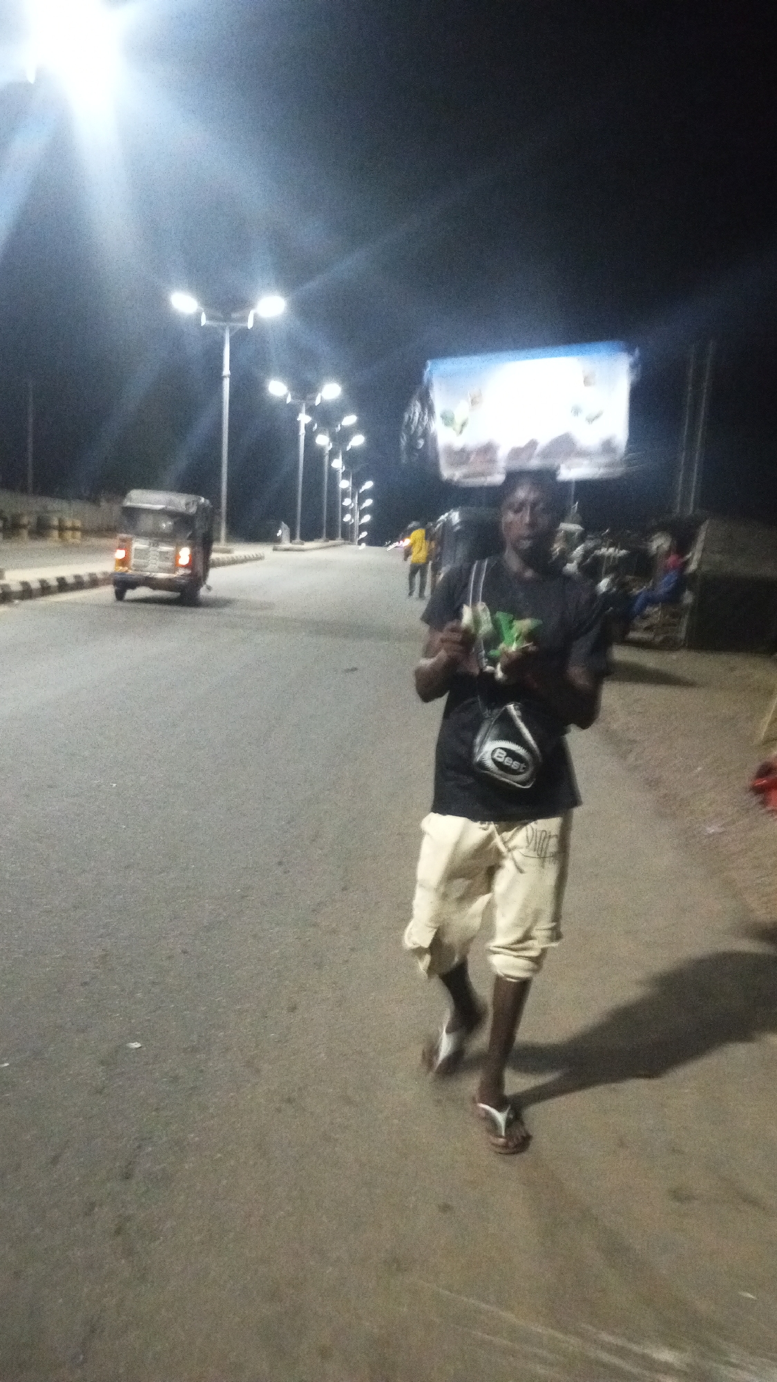 A person carries a container on their head and holds money while walking on a road in Adankolo, Nigeria, at night. The road is lit by streetlights. An auto rickshaw is in the background, and other people are present nearby.