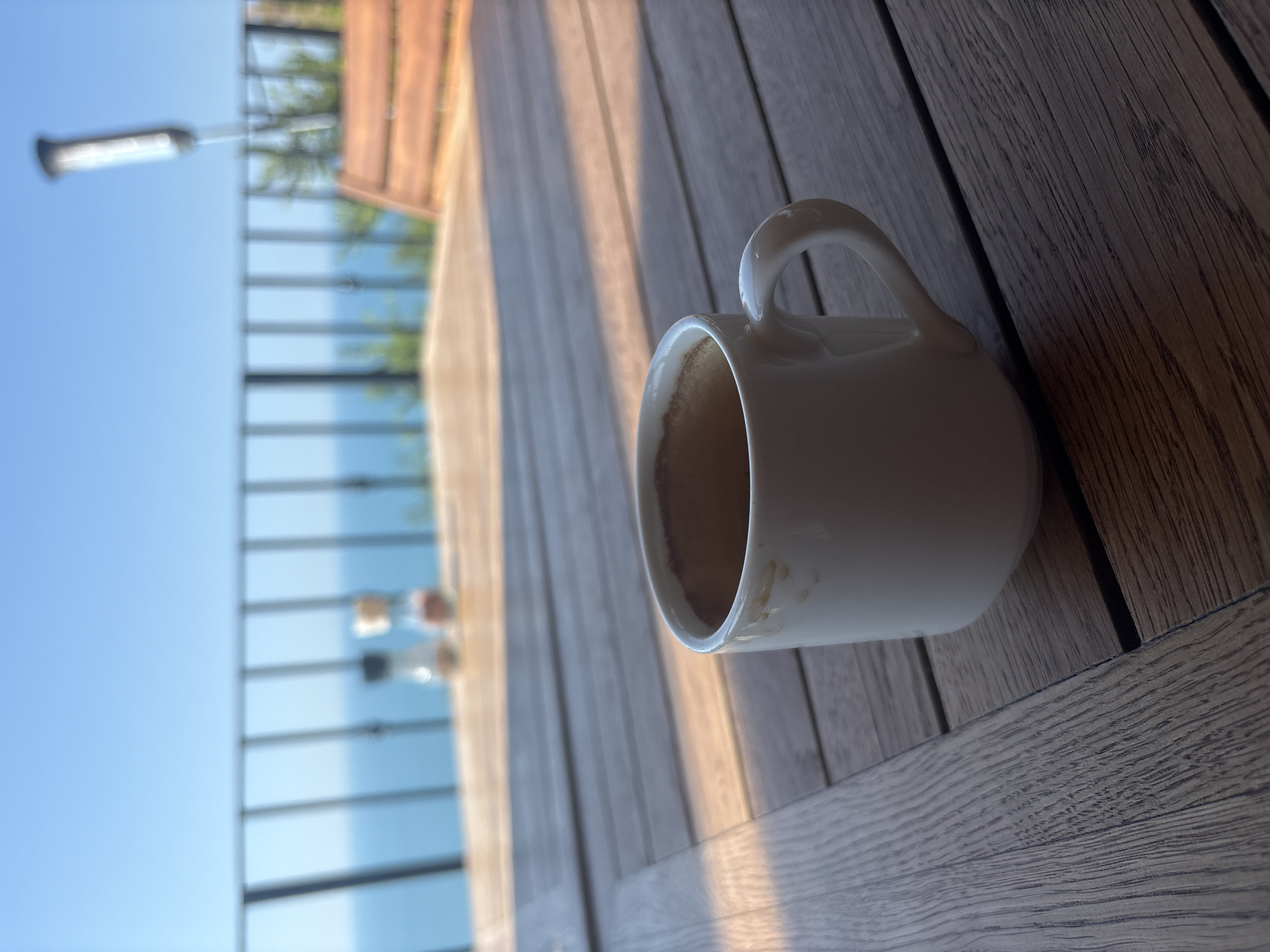 A white ceramic coffee mug, with some brown residue on the inside rim, sits on a wooden outdoor table. The mug is positioned on the right side of the frame, with its handle clearly visible. The table surface is made of wooden planks, with visible grain and texture. In the background, on the left side of the image, a railing with horizontal bars is visible, suggesting an elevated location like a balcony or deck. Beyond the railing, the clear blue sky dominates the upper left portion of the image, while the lower left shows a blurry expanse of blue water. The lighting suggests it is either early morning or late afternoon, with long shadows cast across the table by the sunlight. The overall scene evokes a sense of calm and relaxation, perhaps a moment of quiet contemplation with a cup of coffee enjoyed outdoors. No people are visible in the image, and no text is present.