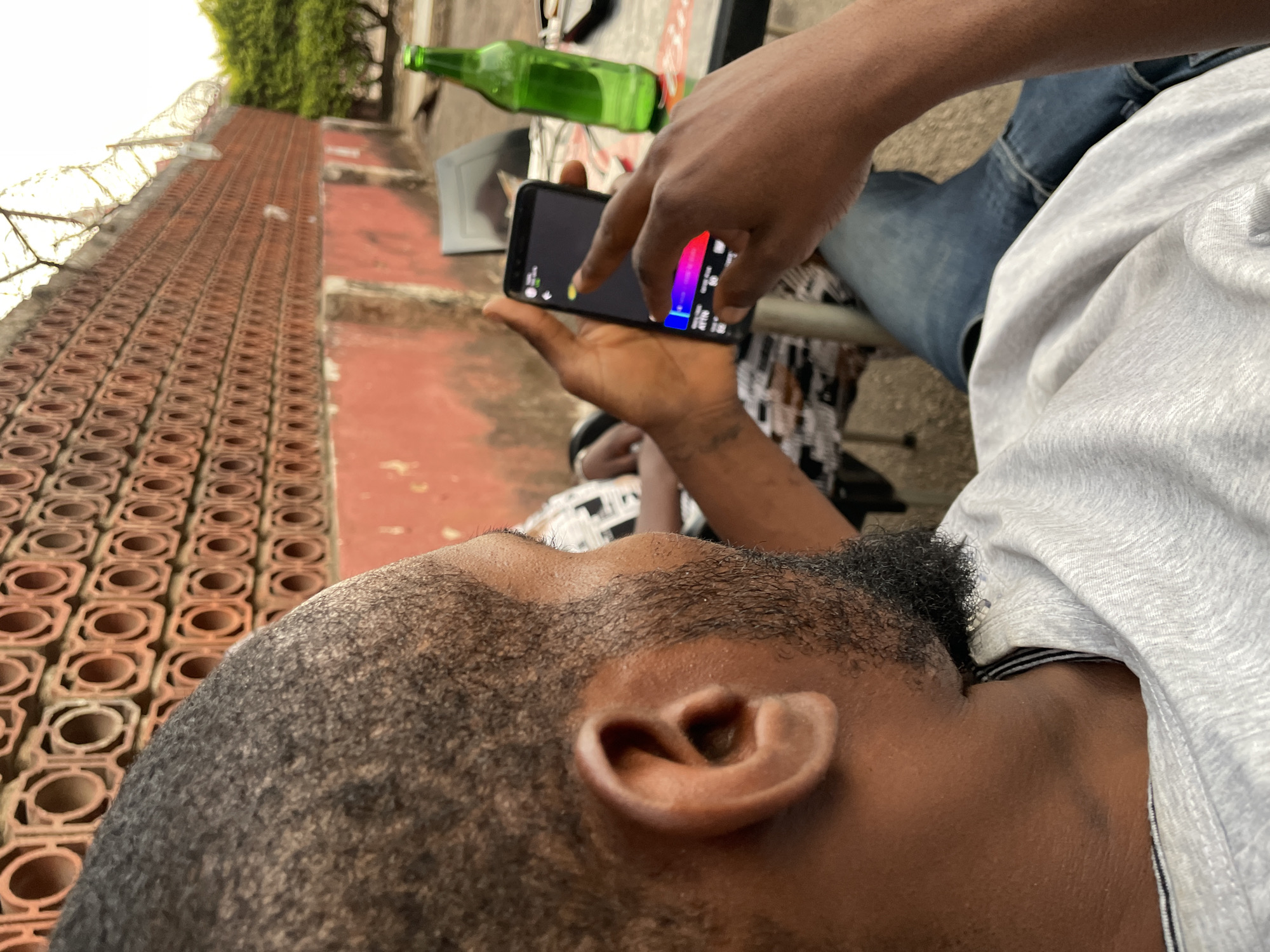 This image, taken in Jos, Nigeria, captures a casual outdoor scene from a high, slightly rotated perspective, looking down.
**Subjects:** The primary subject is a person, likely male, whose head, right ear, and part of his neck and shoulder are visible in the foreground. He has short, dark hair and a beard. His left hand holds a black smartphone, while another hand (possibly his right or a different person's) is touching the screen. In the background, parts of another person are visible, wearing blue jeans and a light grey t-shirt, suggesting they are seated.
**Objects:** The central object is the black smartphone, displaying an interface with a prominent red-to-blue color gradient, along with some smaller icons and text/numbers. A green glass bottle lies out of focus in the mid-ground. The light grey t-shirt and blue jeans are also visible.
**Scene/Setting:** The scene is outdoors, likely a balcony, patio, or rooftop. A distinctive wall or barrier with a repeating square and circular pattern runs along the left side of the frame. The ground beneath appears to be a reddish-orange paved or earthen surface. Some green foliage is visible in the distant background.
**Activity or event taking place:** A person is actively interacting with the smartphone, touching its screen. The interface with the color gradient suggests they might be using a photo or video editing application, a visualizer, or playing a game.
**Notable details:** The lighting indicates it is daytime. The general atmosphere appears relaxed. The camera's angle is tilted, creating a dynamic composition.
**Visible text:** On the smartphone screen, small, indistinct text and numbers are visible, such as "100" and "4770," along with some symbols.