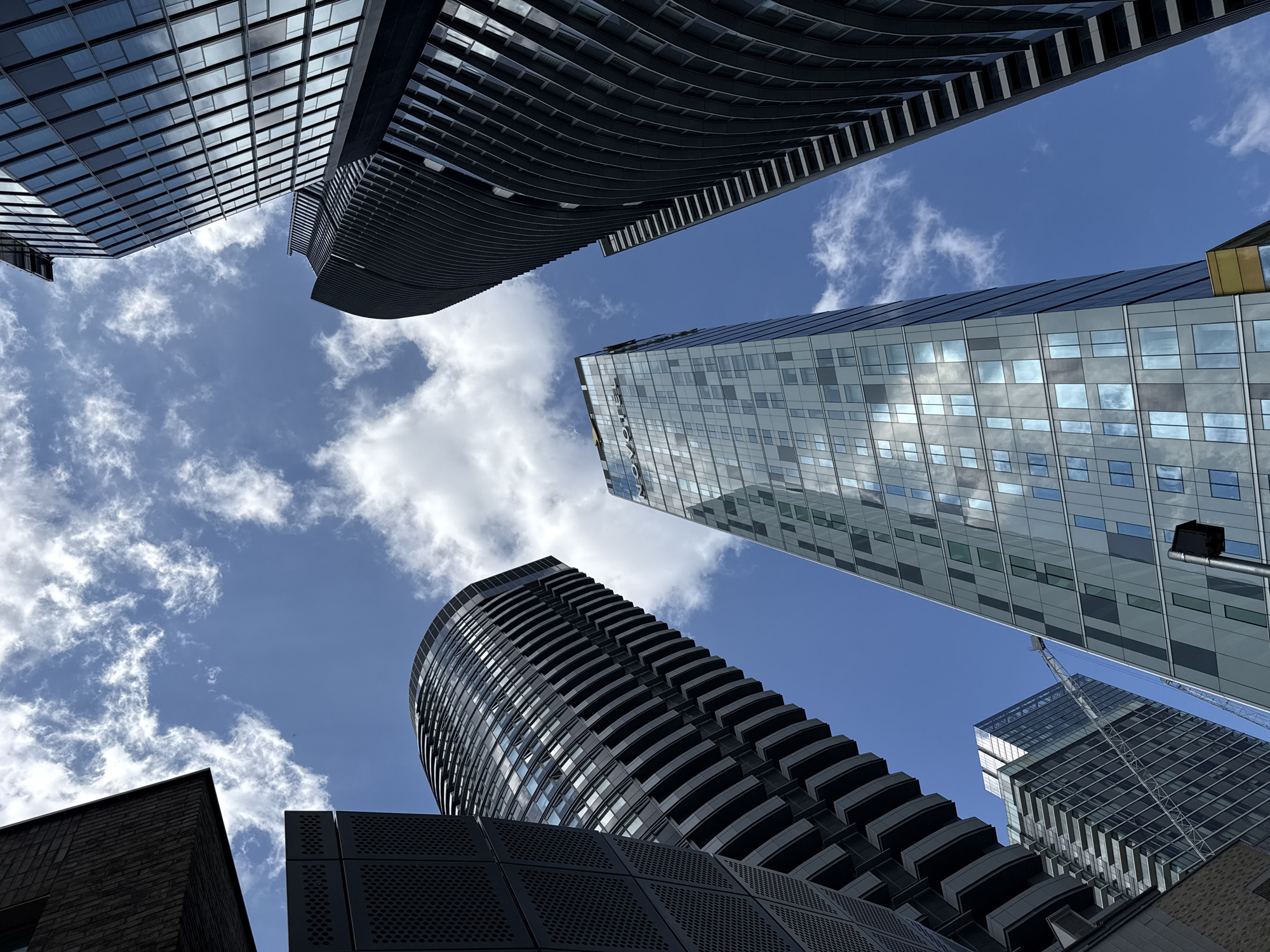This media file captures an upward-looking perspective of several modern high-rise buildings against a clear, vibrant blue sky dotted with scattered white, wispy clouds. The scene is an urban environment, identified as London, United Kingdom, characterized by contemporary architecture.
In the upper left, a building features a rectilinear glass facade that reflects the sky and surrounding structures. Adjacent to it, a dark-colored building with a distinctly undulating or wave-like exterior and horizontal banding or balconies curves upwards.
Towards the right and center, another prominent skyscraper with a mostly glass and steel exterior rises, its numerous windows reflecting patches of blue sky and clouds. Partially visible text, possibly "OVOCA" or similar, is reflected on its surface. A construction crane is visible in the distant background, hinting at ongoing development.
In the lower right, another building with a dark, curved profile and distinct balconies is visible, contributing to the diverse architectural forms. A dark, perforated panel structure is also seen in the lower foreground. The very bottom left corner shows the edge of a brick building, contrasting with the glass and steel towers.
The photograph is taken from a low angle, emphasizing the towering height and scale of the buildings, creating a sense of being enveloped by the urban landscape. The bright sunlight suggests it is daytime, likely mid-day, under fair weather conditions. The composition highlights the verticality and reflective surfaces of modern city architecture.