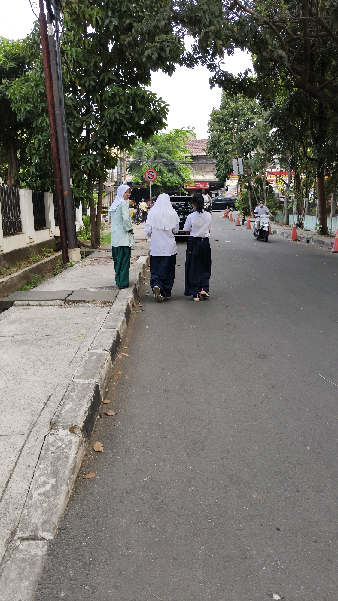 Three young females wearing school uniforms are walking on the asphalt road next to a concrete sidewalk. The female on the far left wears a light green hijab and dark green pants. The two females to her right wear white hijabs and dark blue skirts. They are walking away from the camera, towards a street with traffic. A motorcycle is visible further down the road, with a rider wearing a white shirt and dark helmet. Orange traffic cones are placed along the side of the road. Trees and buildings line the street, suggesting an urban environment. The scene appears to be a typical street in South Jakarta, Indonesia, given the style of architecture and the presence of veiled individuals.