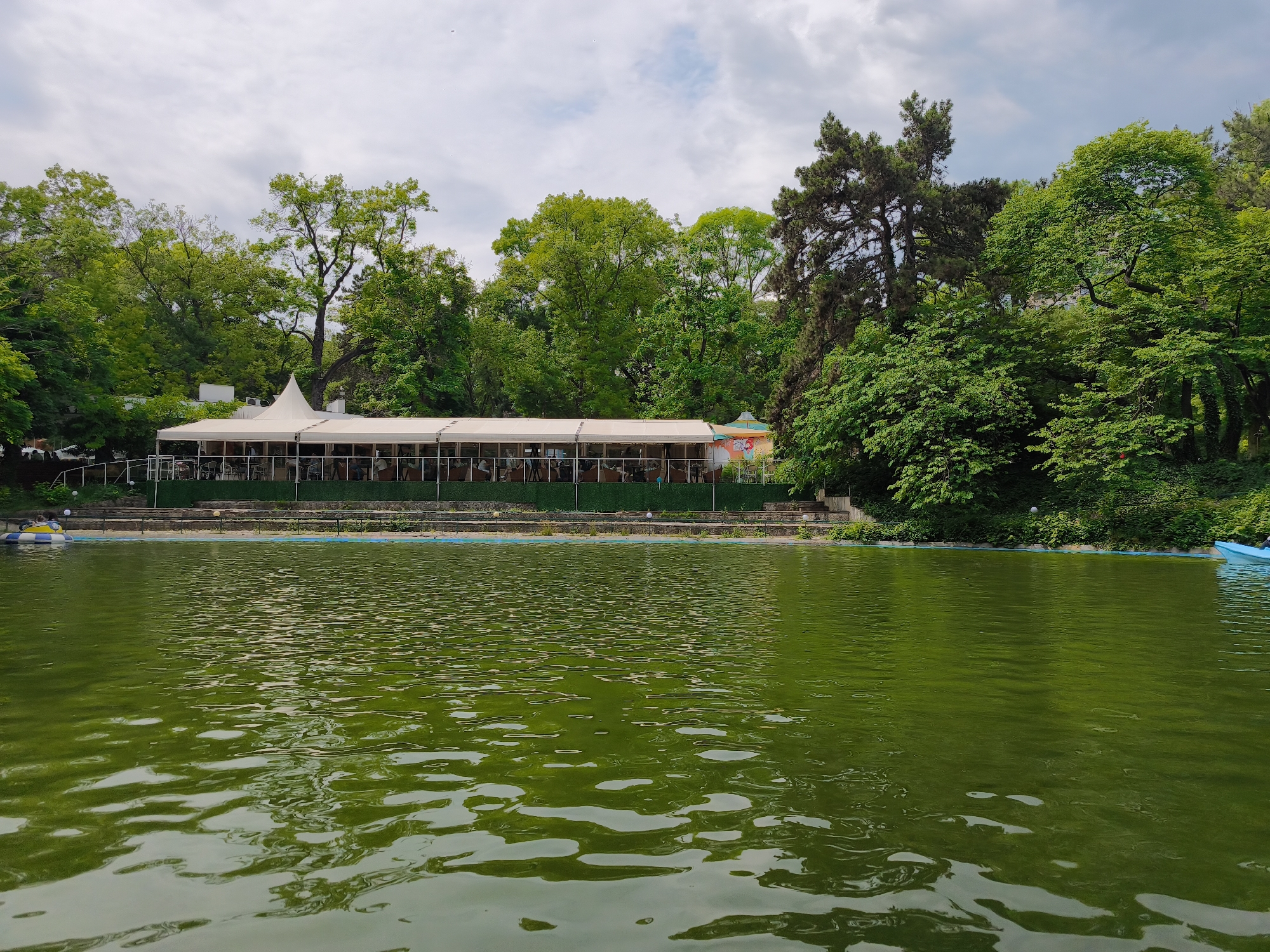 A medium, eye-level shot depicts a green-tinted lake, reflecting an overcast sky, in front of a building and green foliage. The lake occupies the lower two-thirds of the frame, displaying subtle ripples on its surface.
In the background, a long, open-air structure with a white tent-like roof stretches across the frame. People are seated at tables within this structure, enjoying the lakeside view. A green hedge runs along the base of the structure, providing a natural border.
Above the building, a dense canopy of trees fills the upper portion of the image. The foliage is varied, with different shades of green indicating a mix of tree types. The overcast sky provides a soft, diffused light, lending a subdued tone to the scene.
The location is Unknown, Unknown.