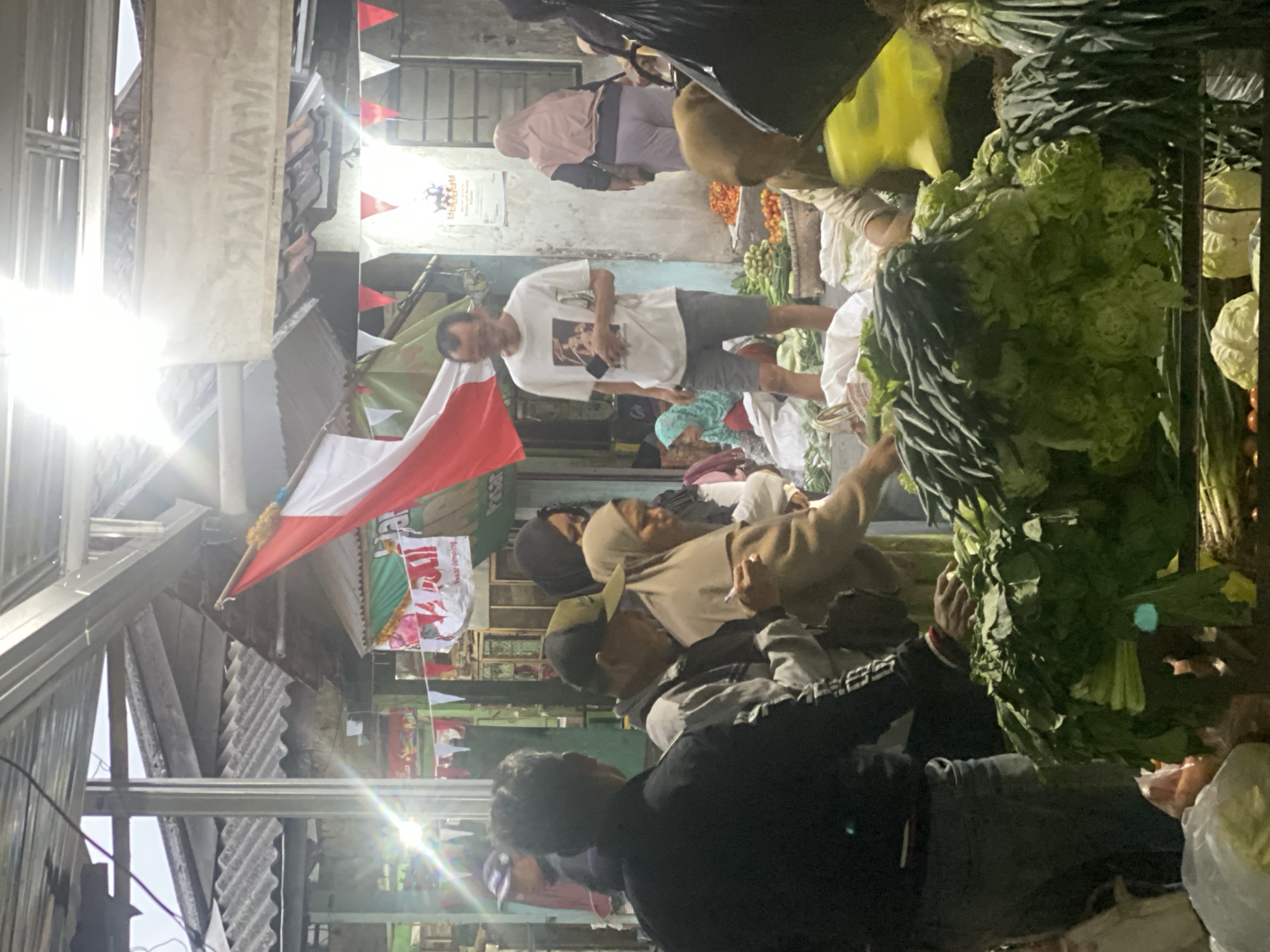 The image depicts an outdoor market scene, possibly in Ambarketawang, Indonesia, given the red and white Indonesian flag prominently displayed. A covered stall structure with corrugated metal roofing and wooden supports is visible overhead. In the foreground, large piles of fresh green vegetables, including lettuce and leafy greens, are arranged for sale. Several people are interacting within the market. A man in a white t-shirt and grey shorts stands near the center, holding a phone. To his left, a man in a black jacket is partially visible. To the right of the man in the white t-shirt, a woman wearing a light brown hijab and a matching scarf is seen. Further in the background, more people and stalls are discernible, with colorful banners and flags adorning the market. The lighting suggests it might be daytime, with the sun casting bright rays through the overhead structure. The overall atmosphere is that of a busy, local marketplace.