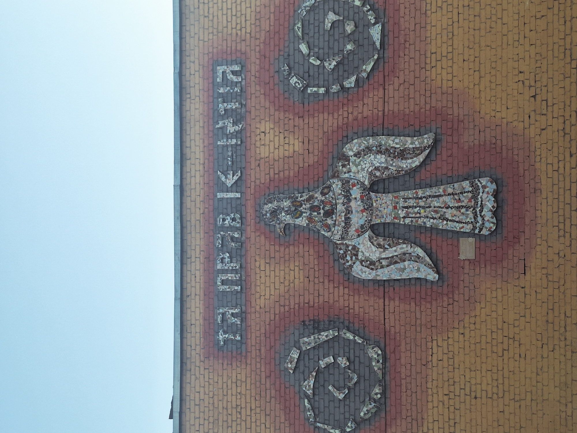 A mosaic on a brick wall depicting a bird-like figure with outstretched wings and swirling patterns above and below it, with Cyrillic text vertically along the side, is situated in an unknown location.