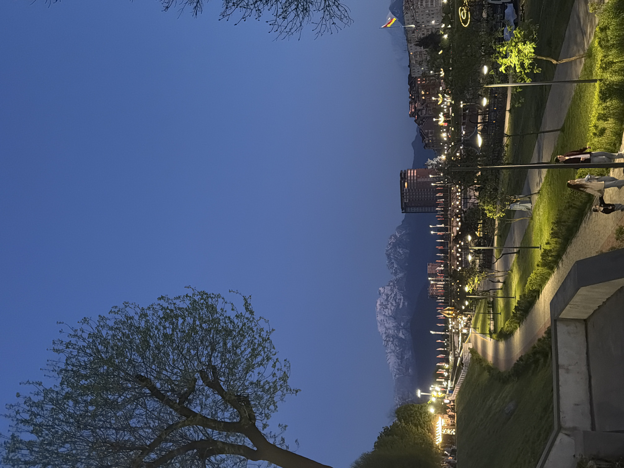 A nighttime, outdoor scene shows a park with lighted pathways in Chermen, Russia. People are walking along the lit paths. Mountains are in the background. The sky is blue. Trees with branches are in the foreground. Buildings are lit in the background. A yellow and red flag is flying.