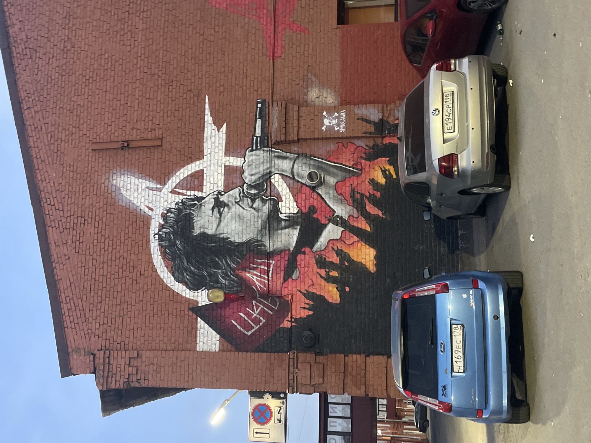 The image shows a mural on a brick building in Saint Petersburg, Russia. The mural depicts a person singing into a microphone, with flames at the bottom. There are two cars parked in front of the building, a blue Ford and a silver Volkswagen. The sky is cloudy. The building is old and has graffiti on it. A street sign with traffic regulations is visible to the left of the mural. The overall scene suggests an urban environment.